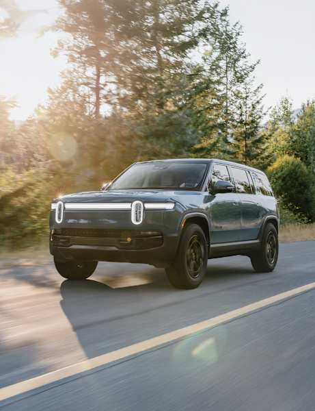 A side-front view of a Green Rivian R1S electric SUV traveling down a paved road, illuminated by bright sunlight filtering through the surrounding forest.