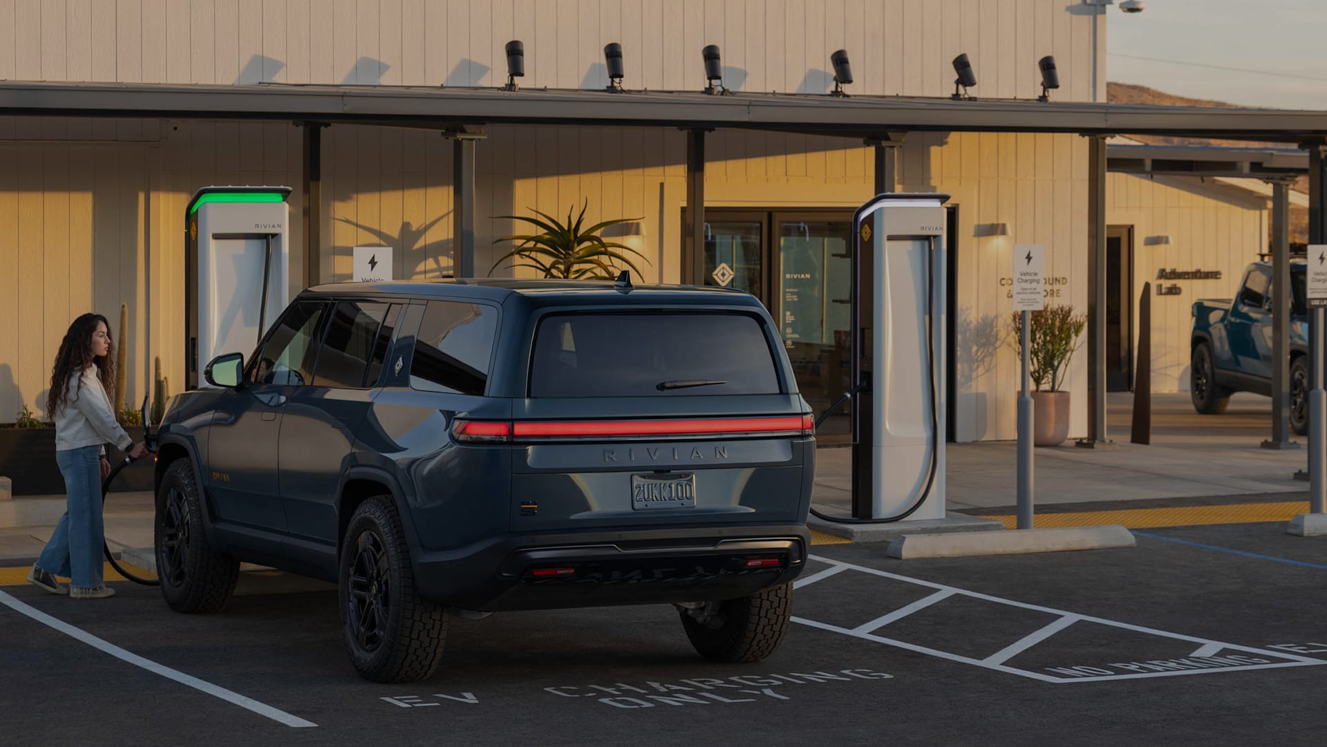 Woman charging a Storm Blue R1S outside at a Rivian space during golden hour.