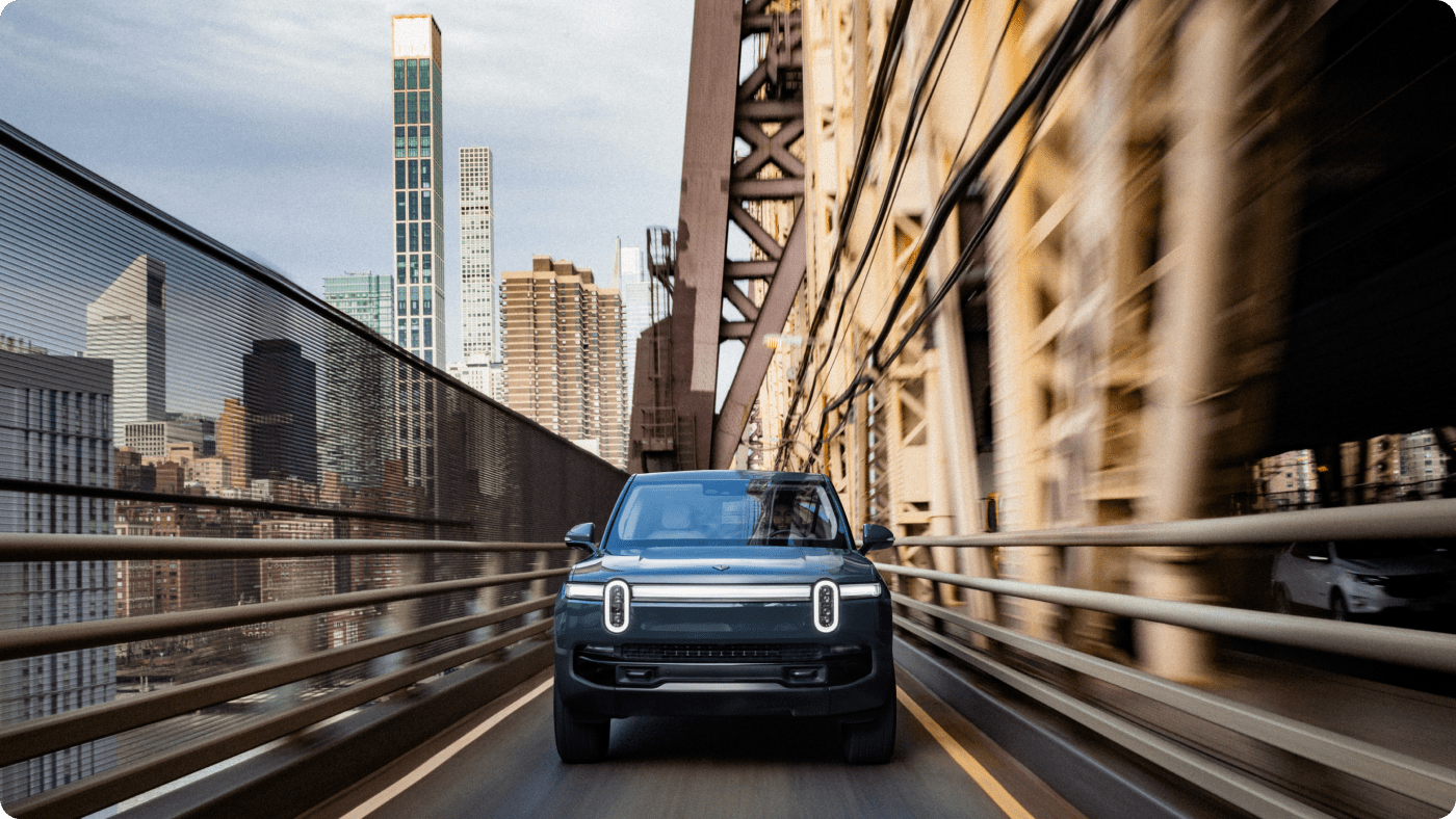 In-motion photo of an El Cap Granite Rivian R1S electric SUV driving over an industrial city bridge.