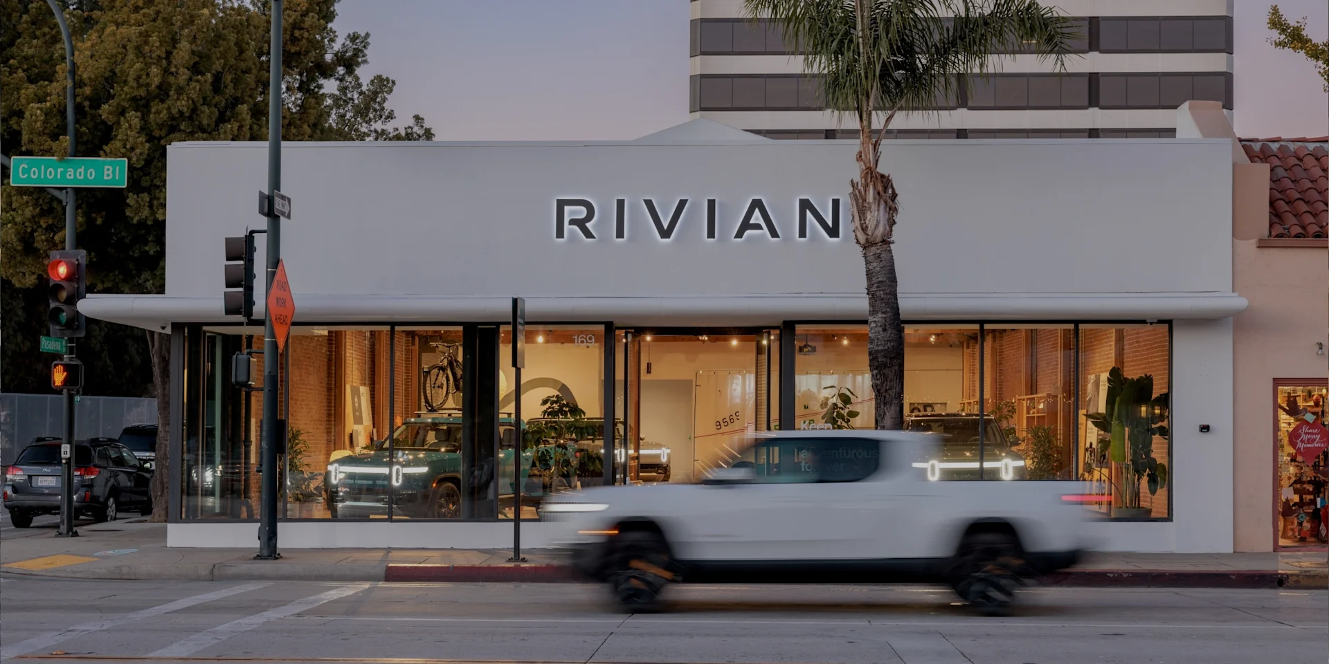 Image of the front of the Rivian Pasadena Space. A white R1T electric truck is moving swiftly on the street in front of the store.