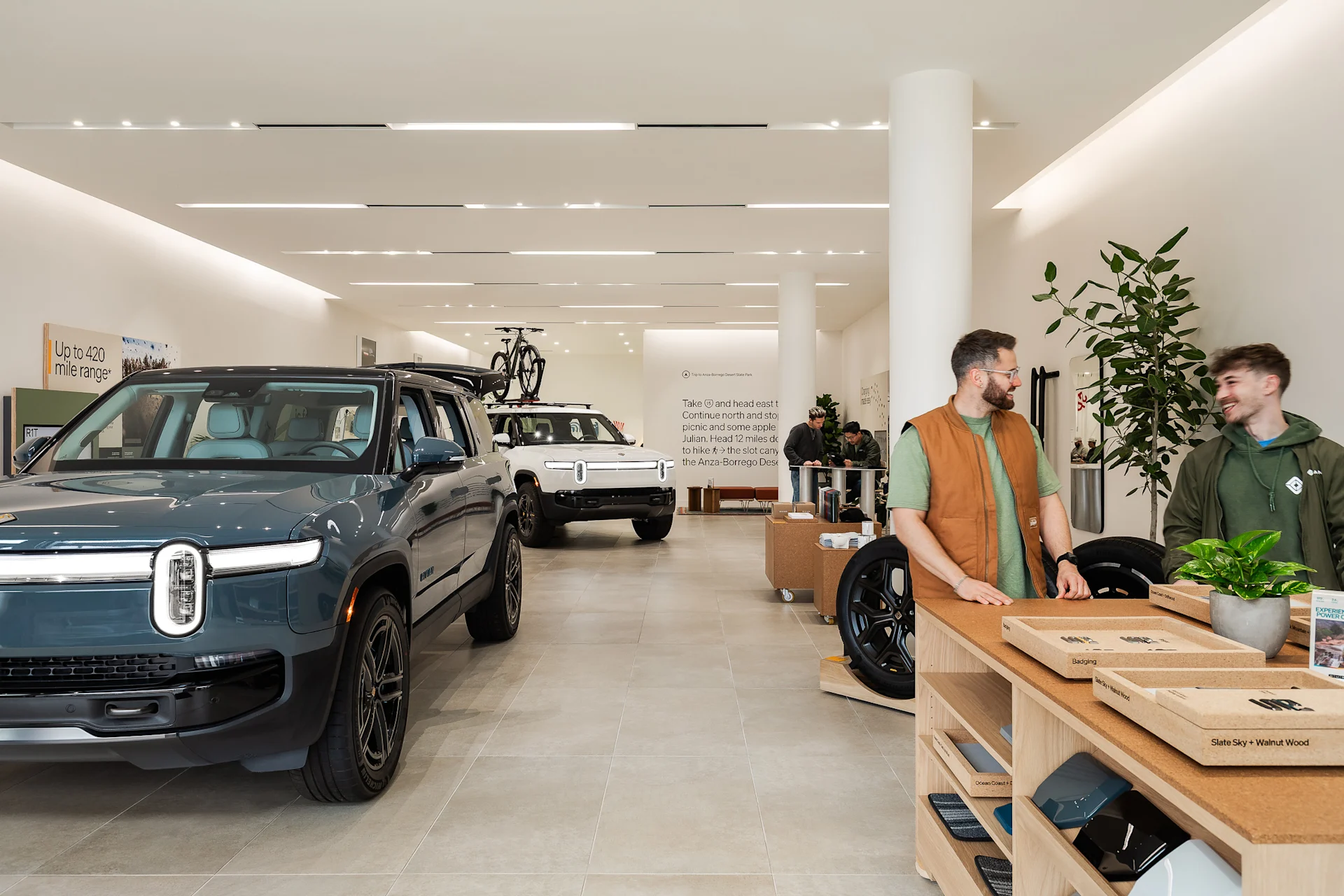 The inside of the Rivian La Jolla space. A dark blue Rivian R1S and and white Rivian electric vehicle are parked in a link, while an advisor chats with a customer.