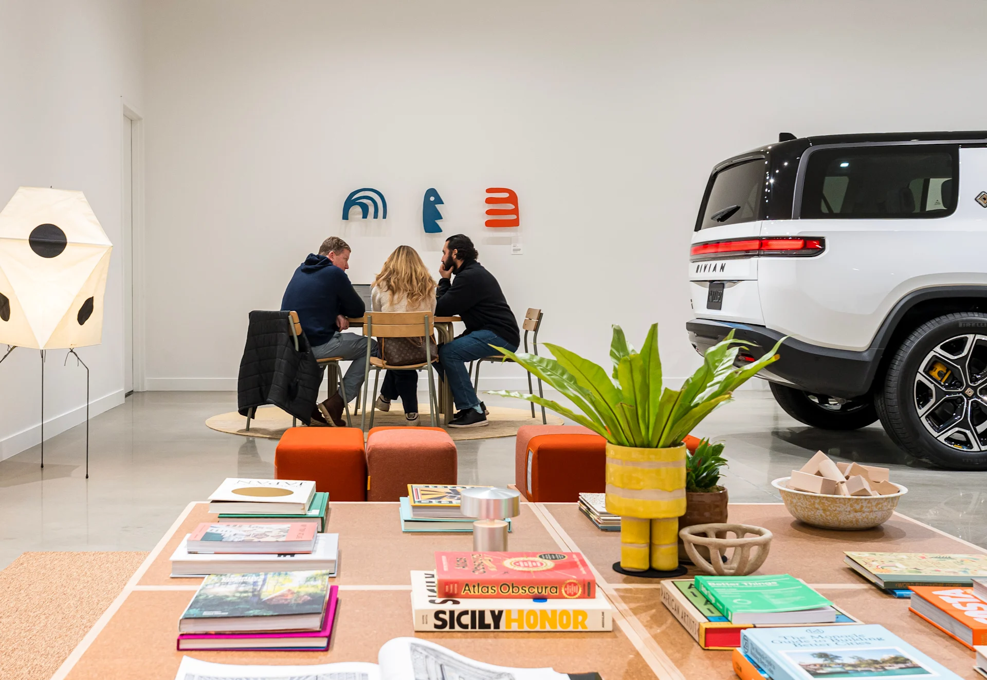 Three people sitting at a table beside a white Rivian R1S electric SUV inside the Rivian King of Prussia space.