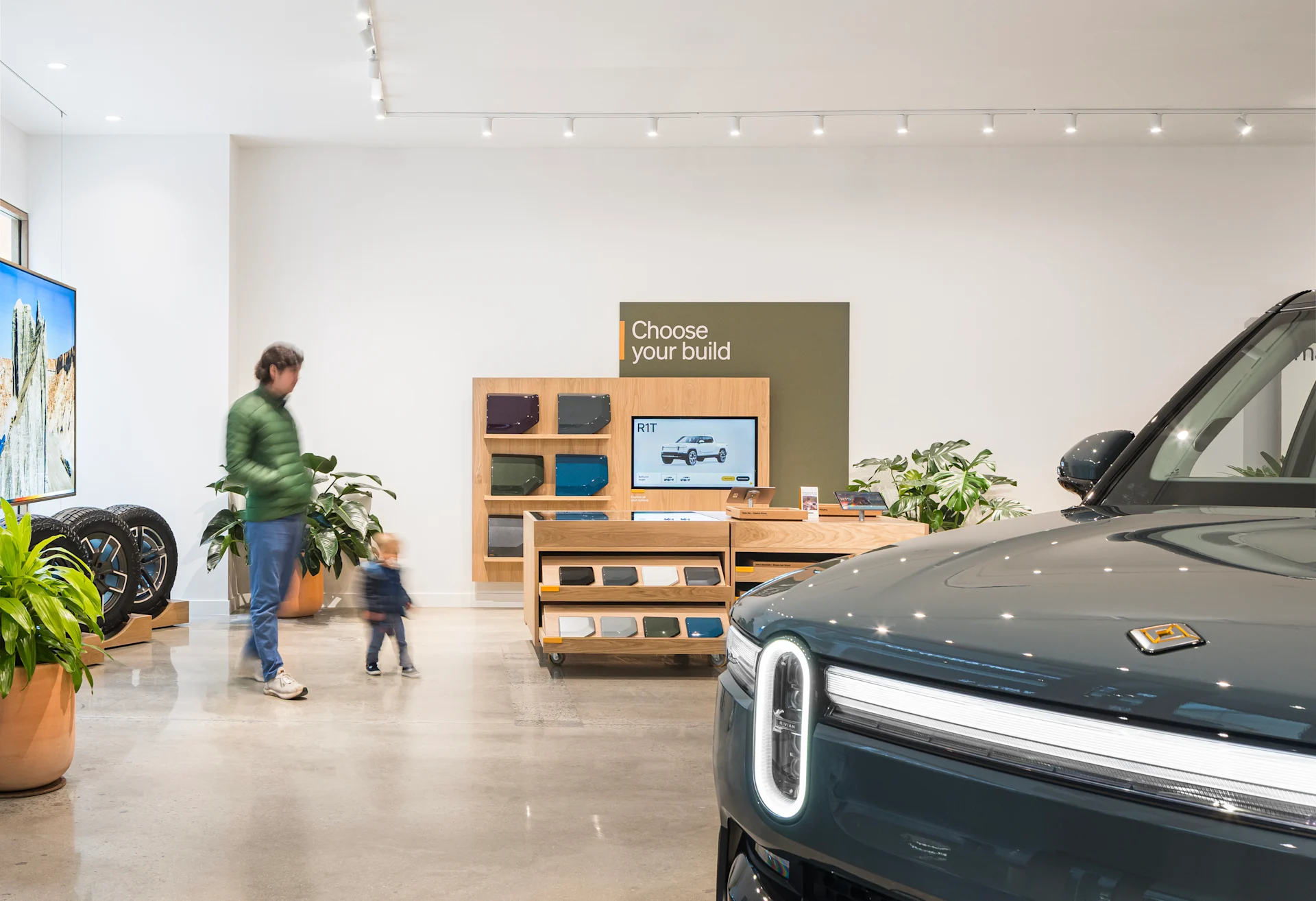 A father and child walking through the Rivian Rockling Service + Demo Center. A dark blue Rivian EV SUV takes up the bottom right corner.