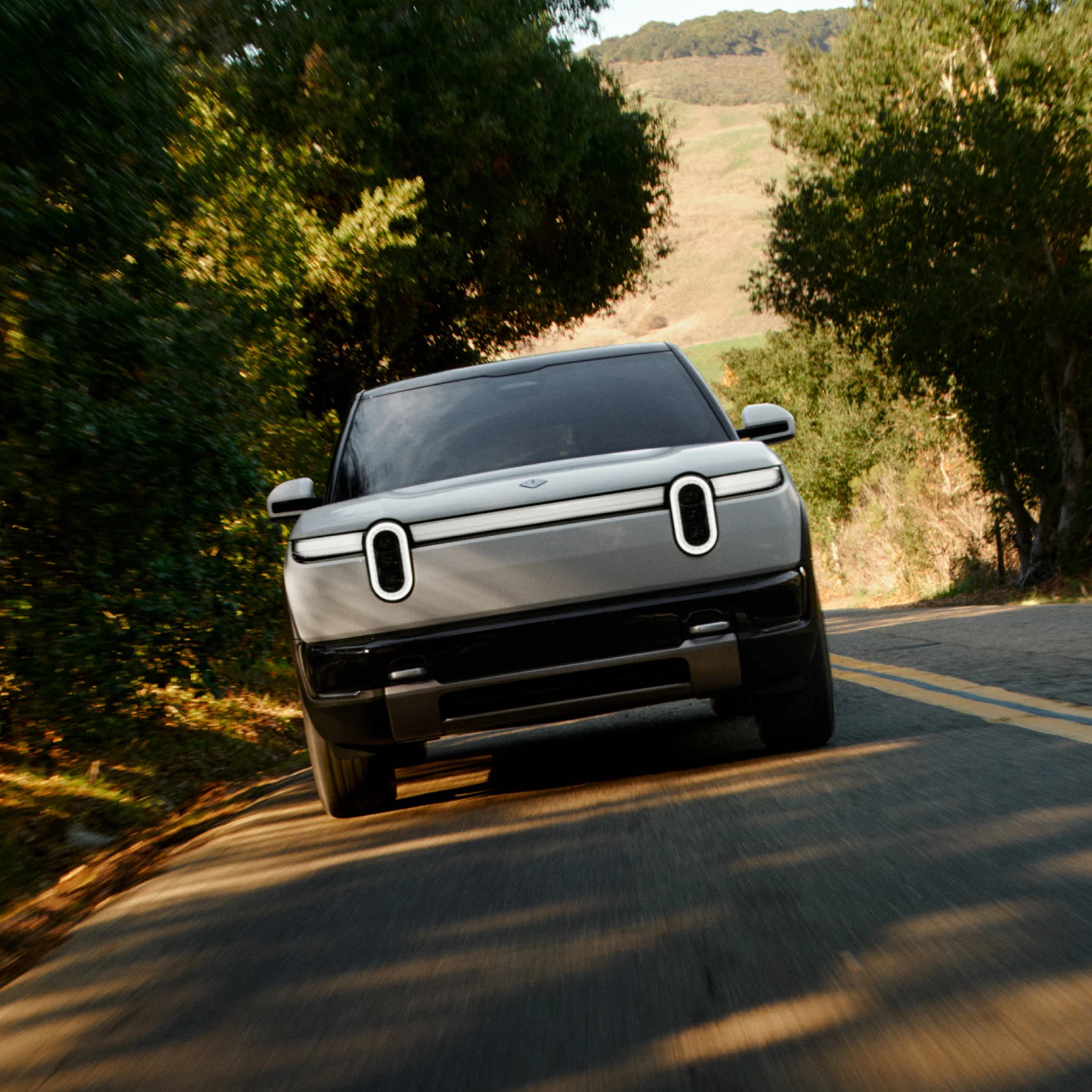 Front view of a white Rivian R2 electric SUV driving on a winding, tree-lined road. The SUV features distinctive stadium headlights and a full-width light bar. Shadows stretch across the asphalt from surrounding trees.