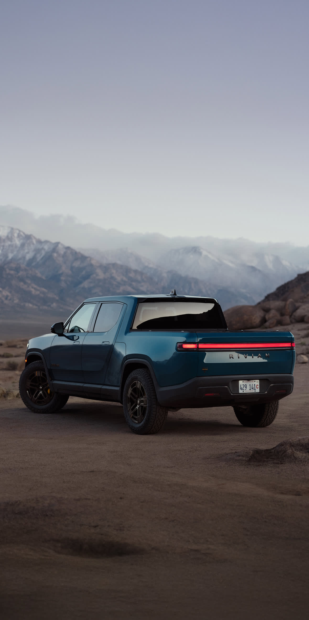 A blue Rivian R1T electric truck is parked on a dirt path in a vast, open desert landscape. In the background, rugged mountains with snow-covered peaks sit beneath a soft, hazy sky at dusk.