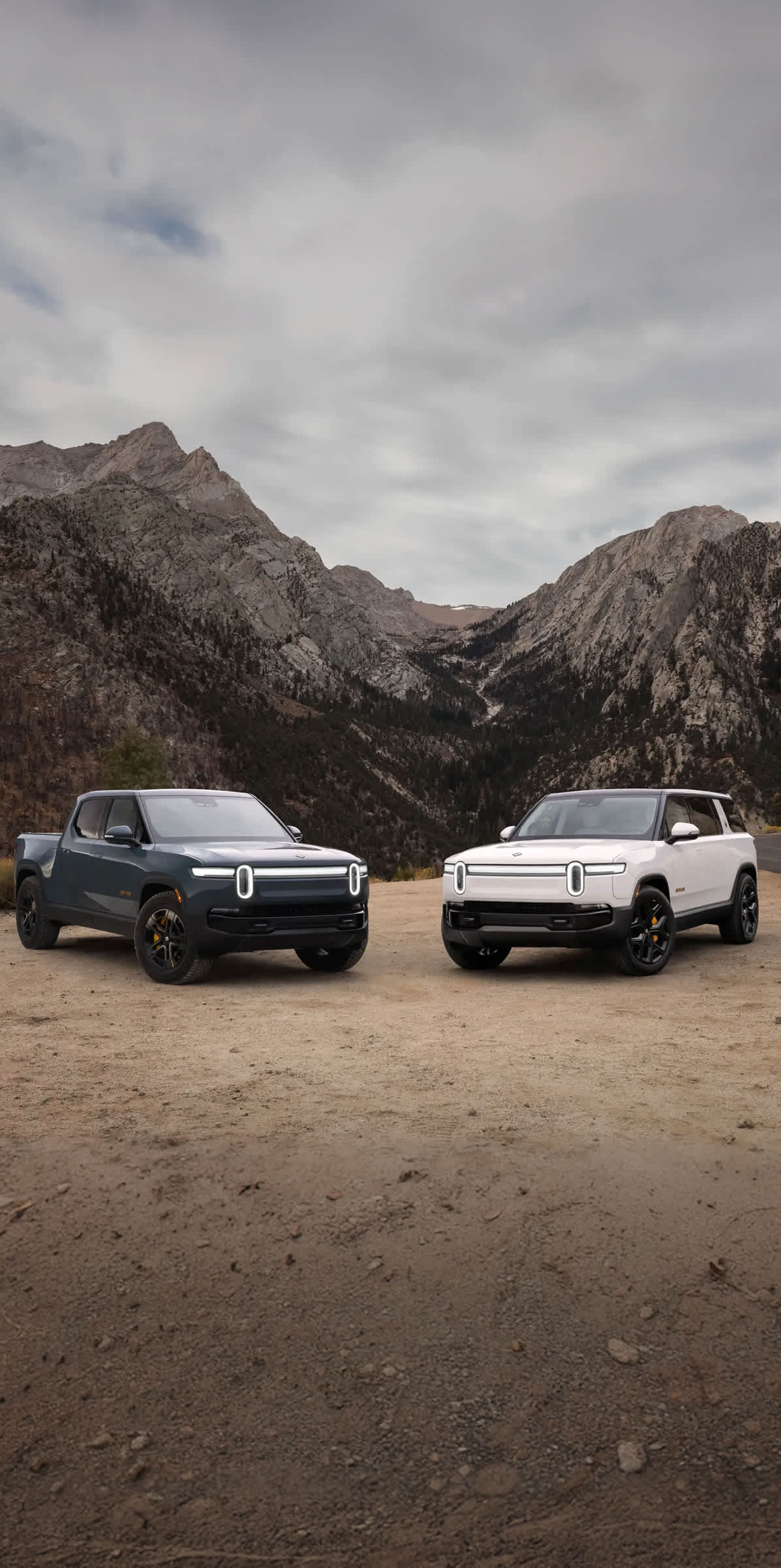 A Rivian R1S electric SUV and R1T electric truck, parked on a dusty side road with scenic desert mountains in the background.