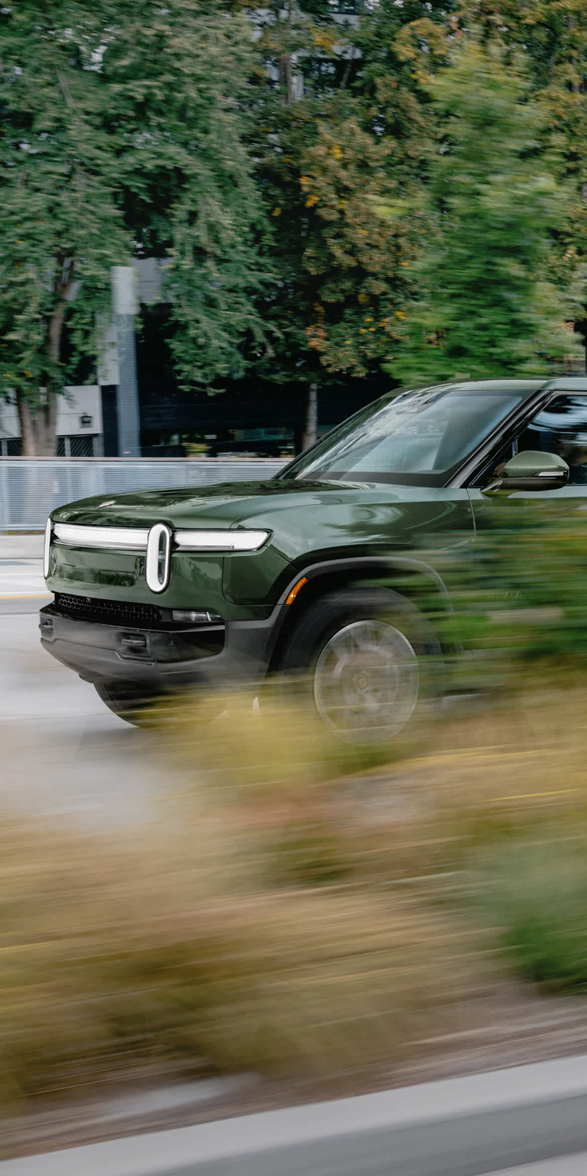 A forest green Rivian R1T electric pickup truck driving on a paved city street. The image uses a motion blur effect on the foreground foliage and background trees to emphasize speed.
