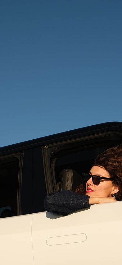 A side-angle view of the White Rivian R2 Electric SUV featuring a passenger leaning out of the front window with her hair blowing in the wind
