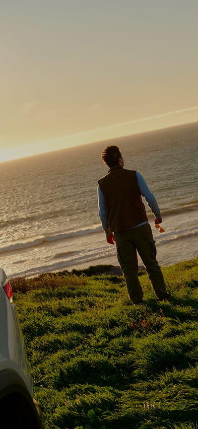 A low-angle, wide shot of a man and a woman standing on a lush green grassy hill at sunset, looking out toward the ocean. To the left, the rear of a white R2 Rivian electric SUV is visible with its trunk open.