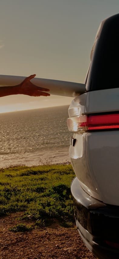 A person slides a surfboard through the rear window of a Rivian R2 electric SUV parked on a grassy cliff at sunset.