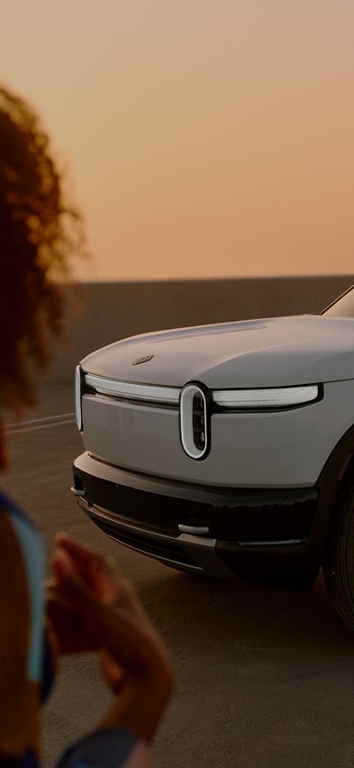 Front three-quarter view of a white Rivian R2 electric SUV at sunset, featuring the signature light bar and stadium headlights with silhouettes of people in the foreground.