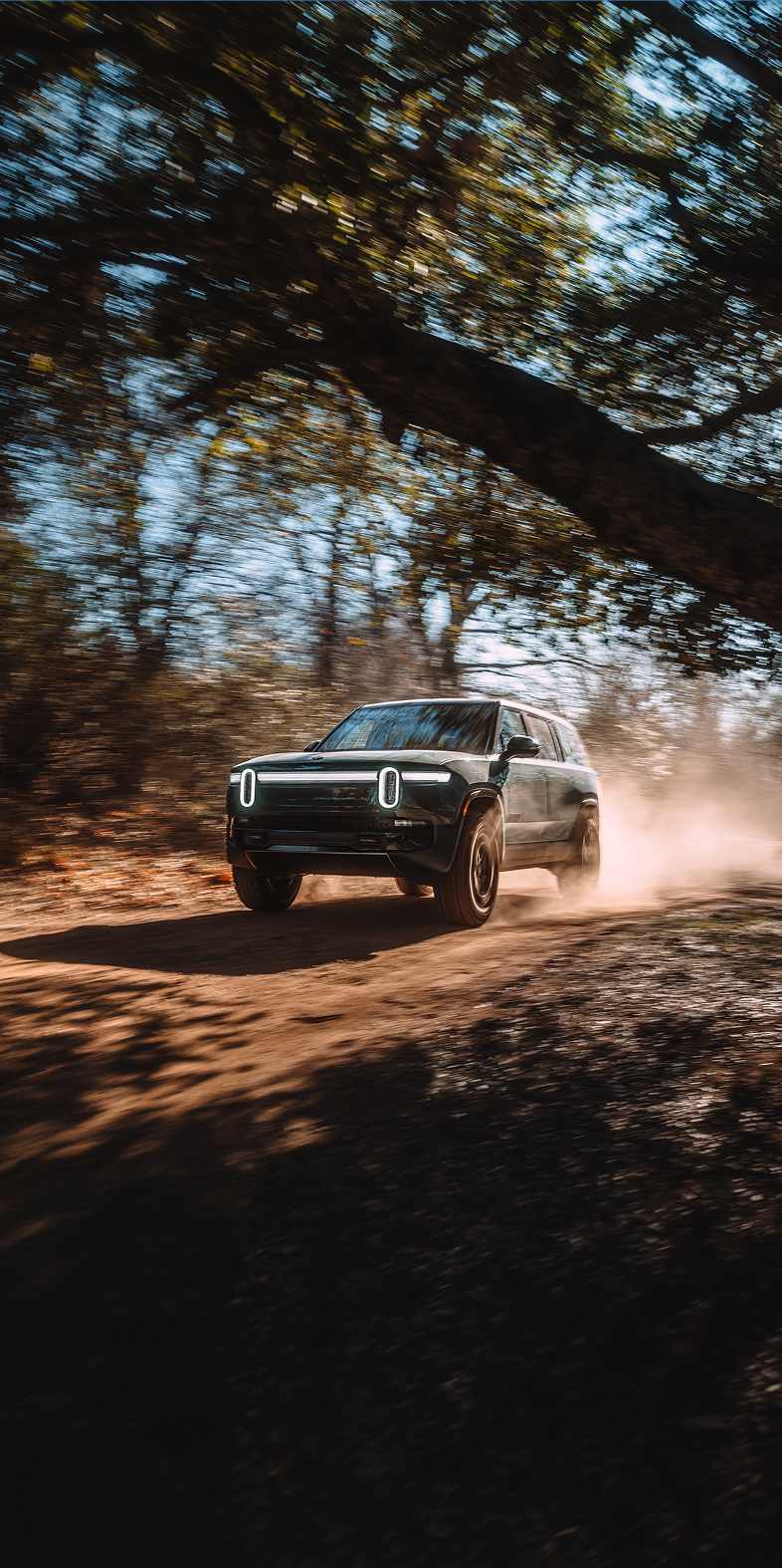 A dark green Rivian R1S electric SUV driving along a dirt path in a forest, kicking up a cloud of dust.