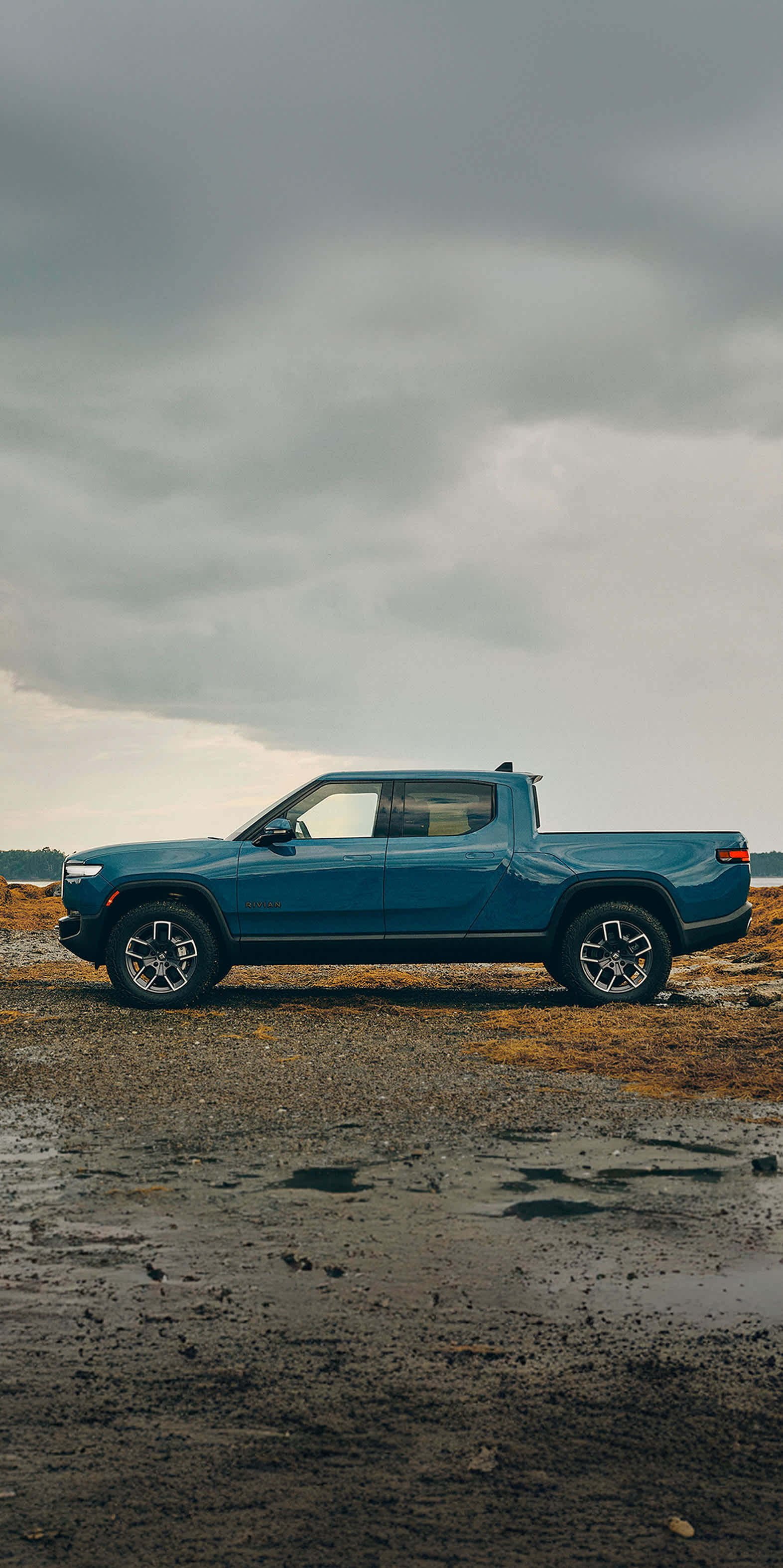 A rugged blue Rivian R1T electric truck in front of a scenic rocky shore of a lake. 