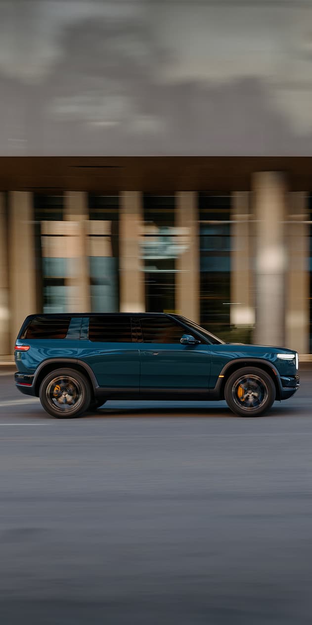A Catalina Blue Rivian R2 electric SUV driving past blurred city buildings, captured in a side-profile motion shot with a black roof and yellow brake calipers.