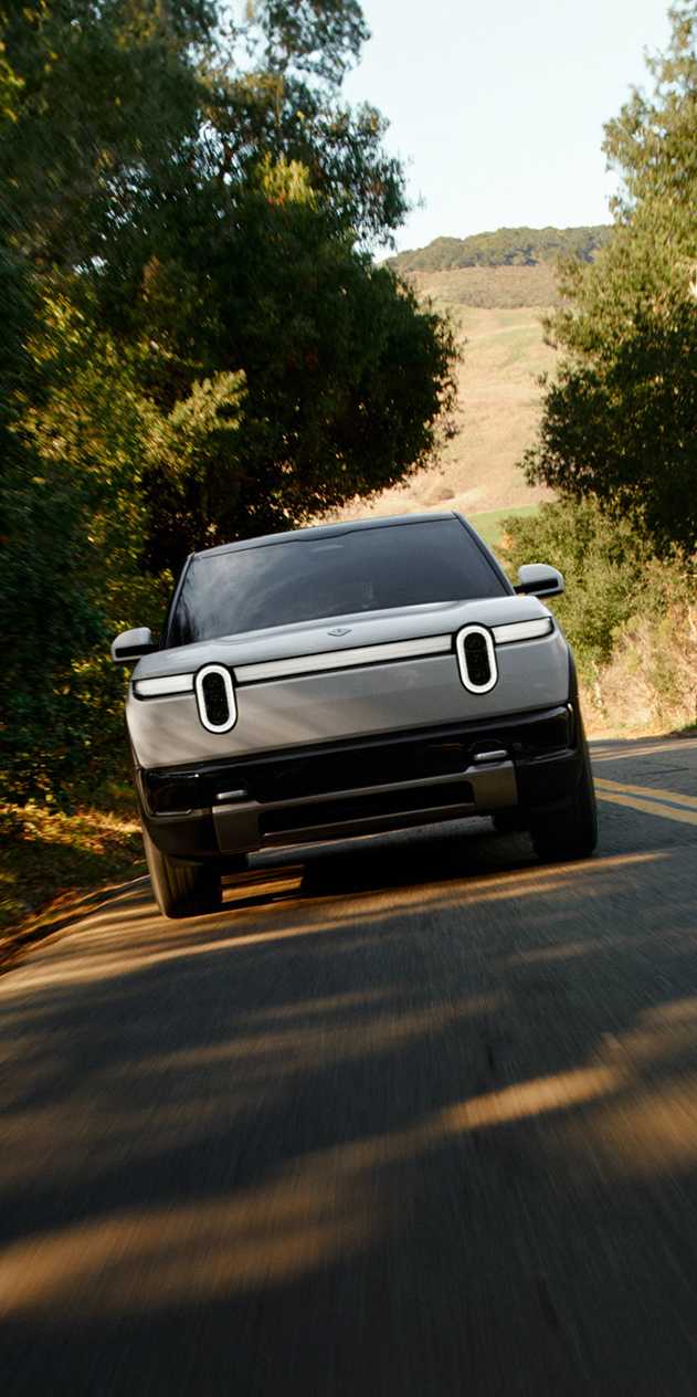 Front view of a white Rivian R2 electric SUV driving on a winding, tree-lined road. The SUV features distinctive stadium headlights and a full-width light bar. Shadows stretch across the asphalt from surrounding trees.