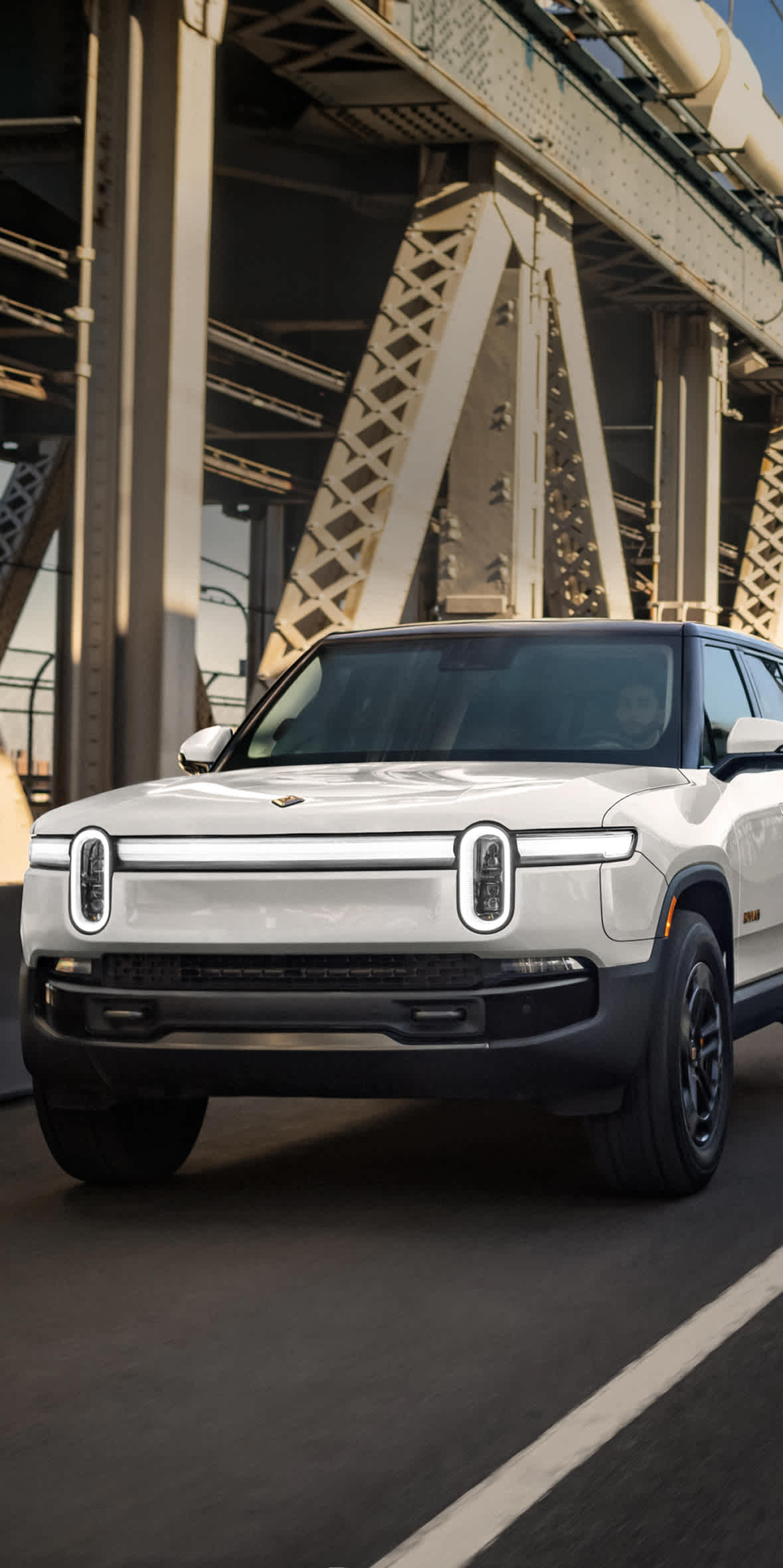 A white Rivian R1S electric SUV drives across a Bridge, showing that rugged capability looks just as good against a city skyline as it does on a mountain trail.