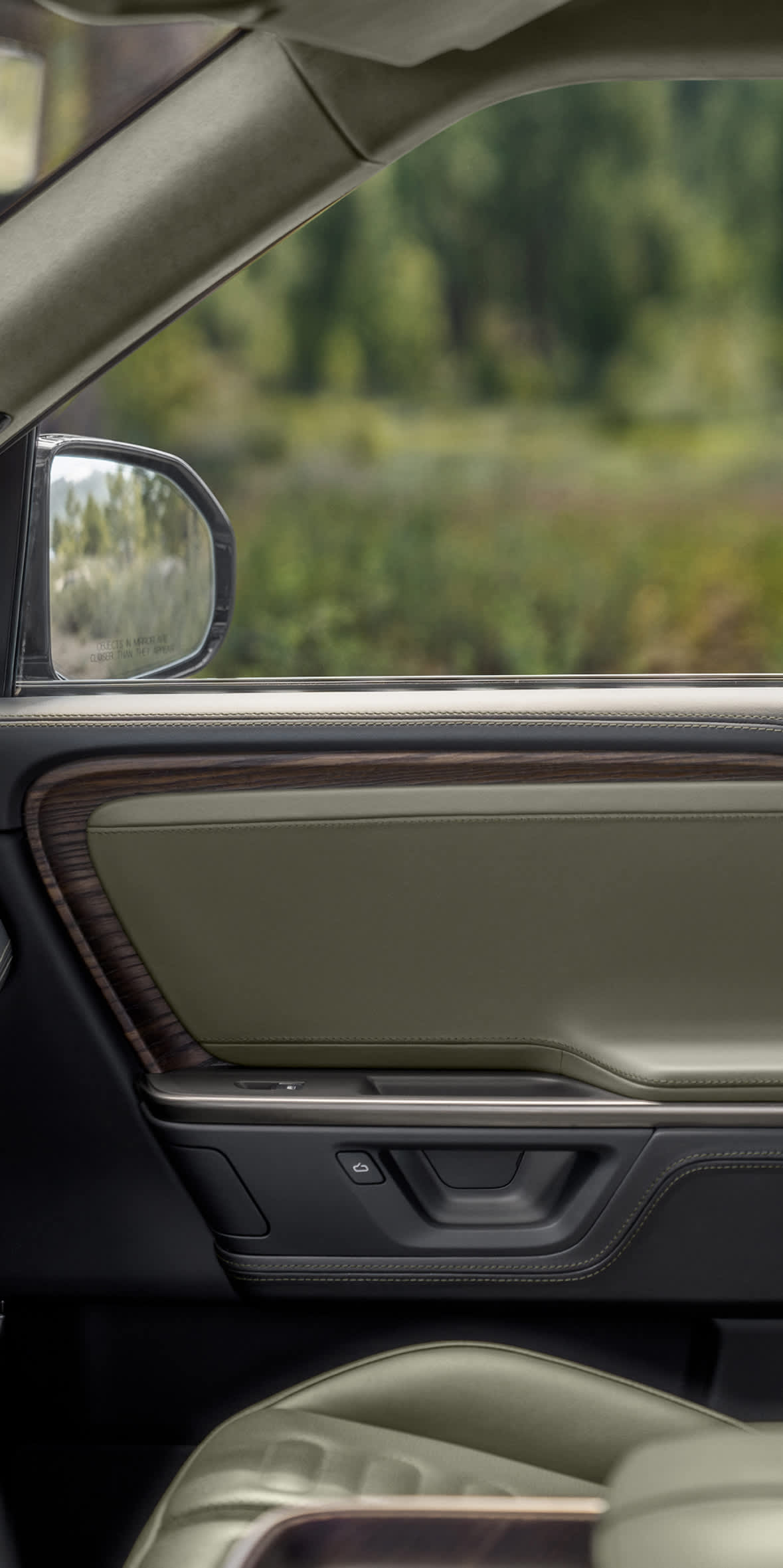 Interior view of an electric Rivian R1 SUV featuring the Forest Edge Package, showcasing olive green upholstery, warm wood accents on the door panel, and a view of lush green trees through the passenger window.