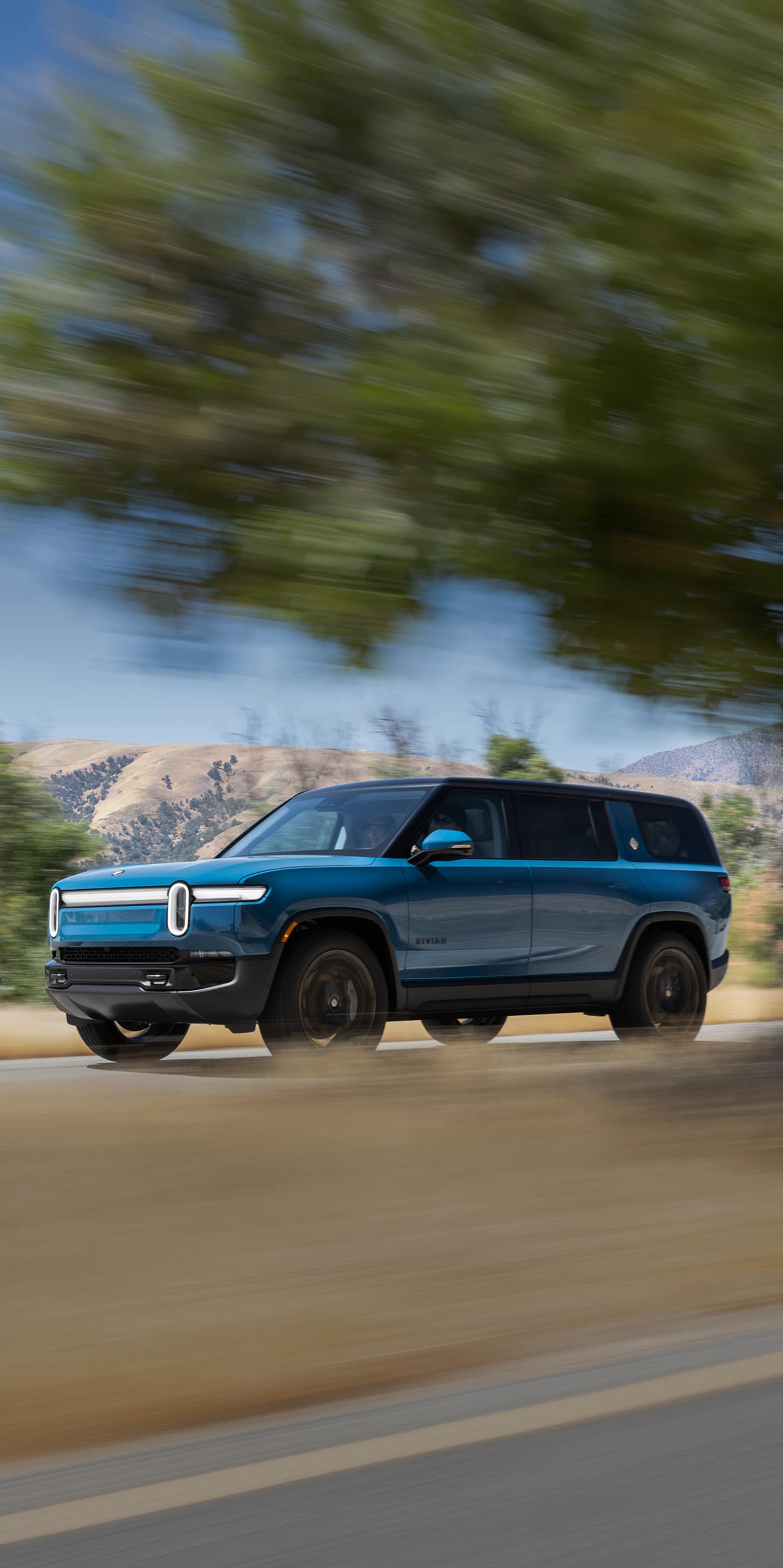 A Rivian Blue R1S electric SUV driving swiftly down an open highway. Mountains and bright blue skies fill the background.