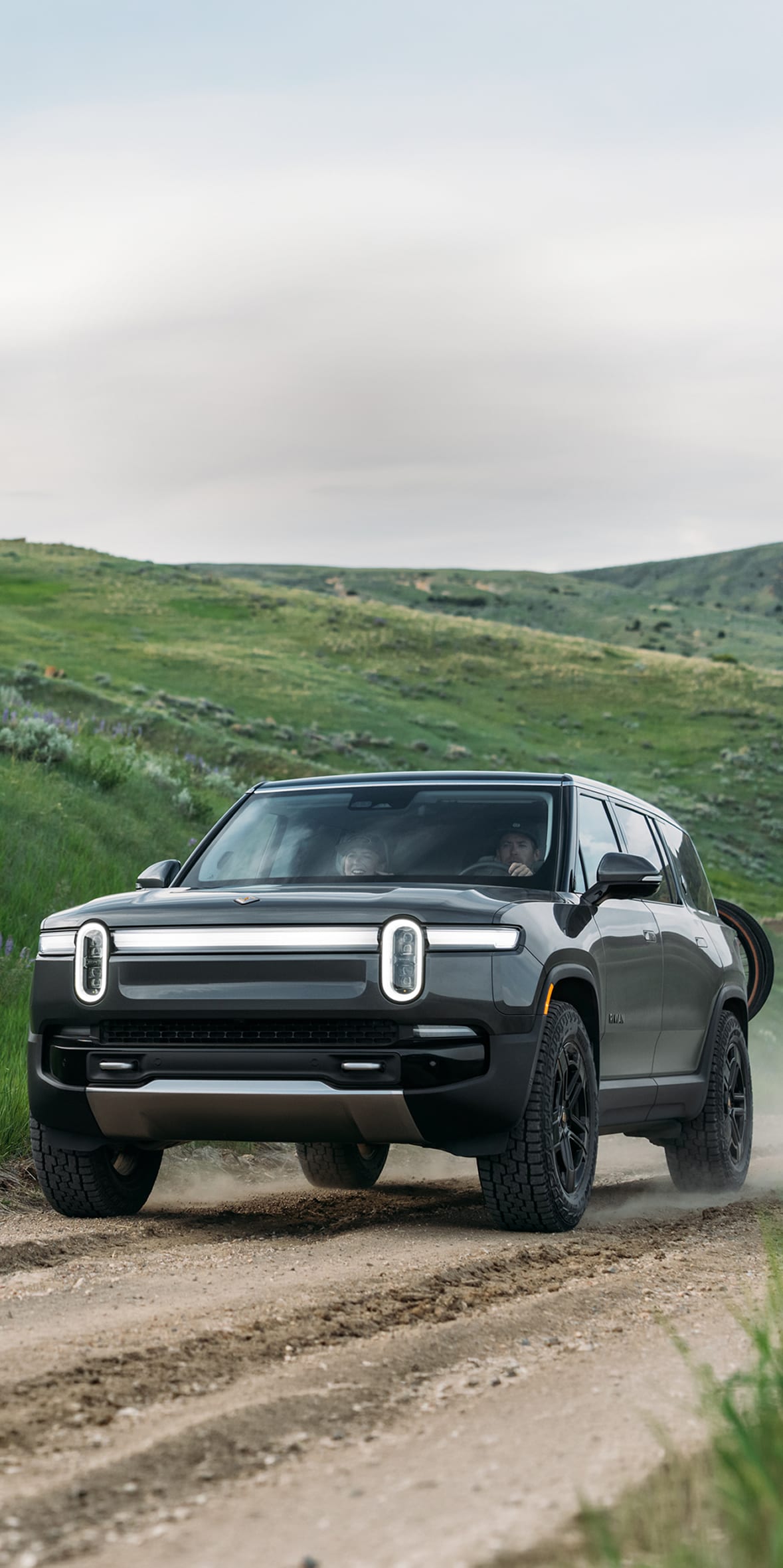 Front view of a dark grey Rivian R1S electric SUV driving along a dirt road, kicked-up dust visible behind its wheels, with vibrant, rolling green hills in the background.