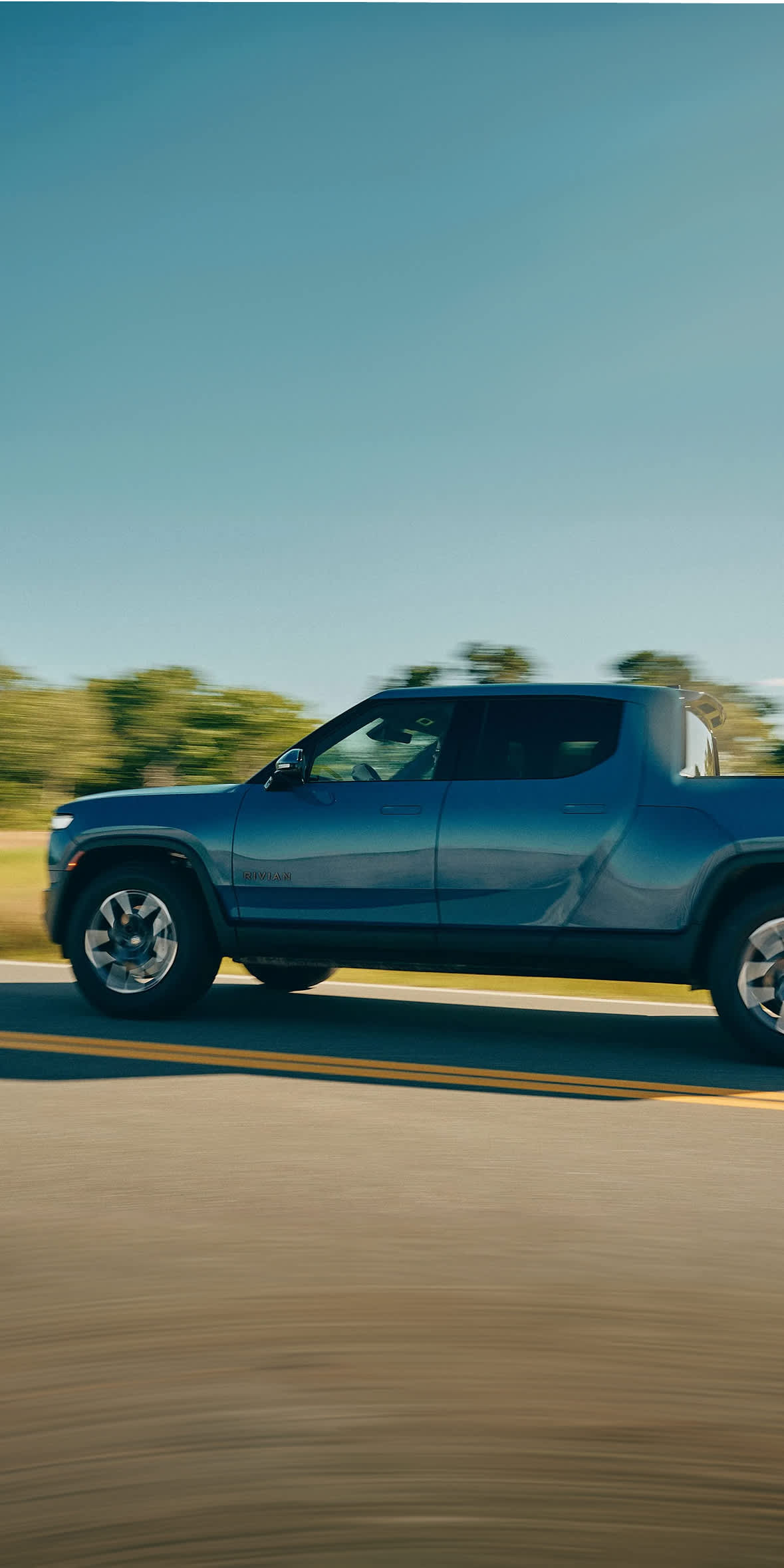 An all-electric Rivian R1T pickup truck in Rivian Blue cruises along a two-lane asphalt road with yellow lines. The side profile reveals a motion-blur background of green trees and grass under a clear blue sky, emphasizing speed.