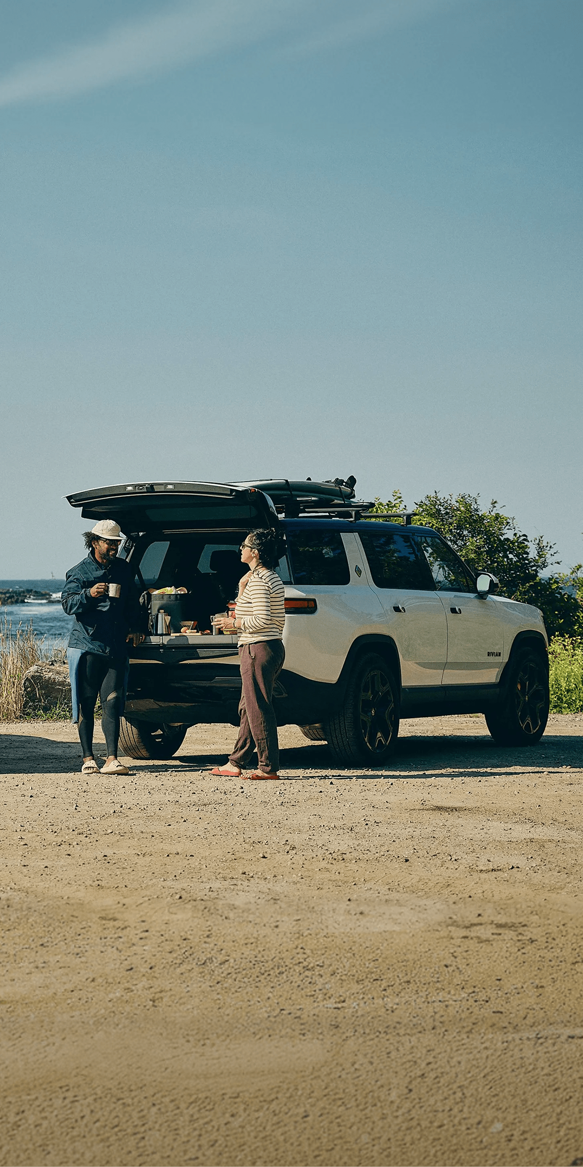 A blue R1T electric truck and a white R1S electric SUV parked by a rocky coastline. Three people are socializing in the sunshine near the open tailgate of the white SUV.
