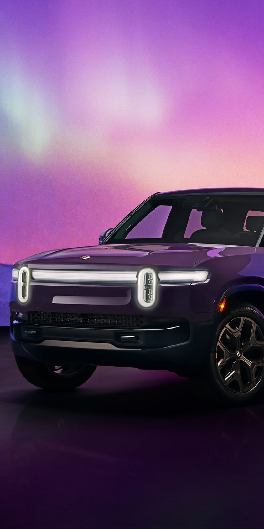 A side-front view of a Borealis purple Rivian R1S electric SUV in a stylized studio setting. The R1S is positioned on a dark surface, illuminated against a vibrant purple and magenta background evoking the colors of the Aurora Borealis.
