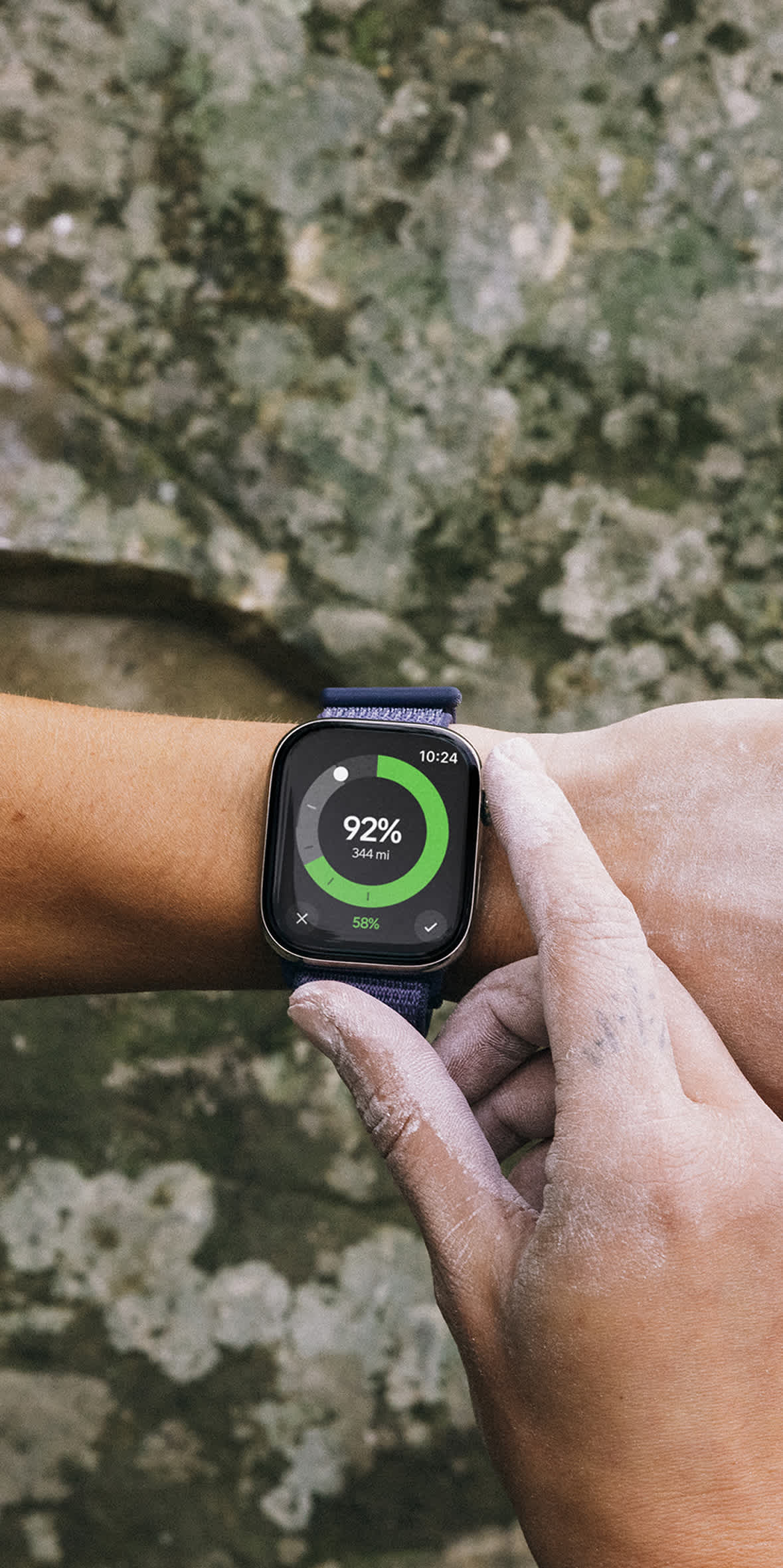 An over-the-shoulder view of a climber with chalk-covered hands looking at an Apple Watch shown on a wrist with the Rivian app charging screen. The watch displays a 92% charge and 344 miles of range against a textured rock background.
