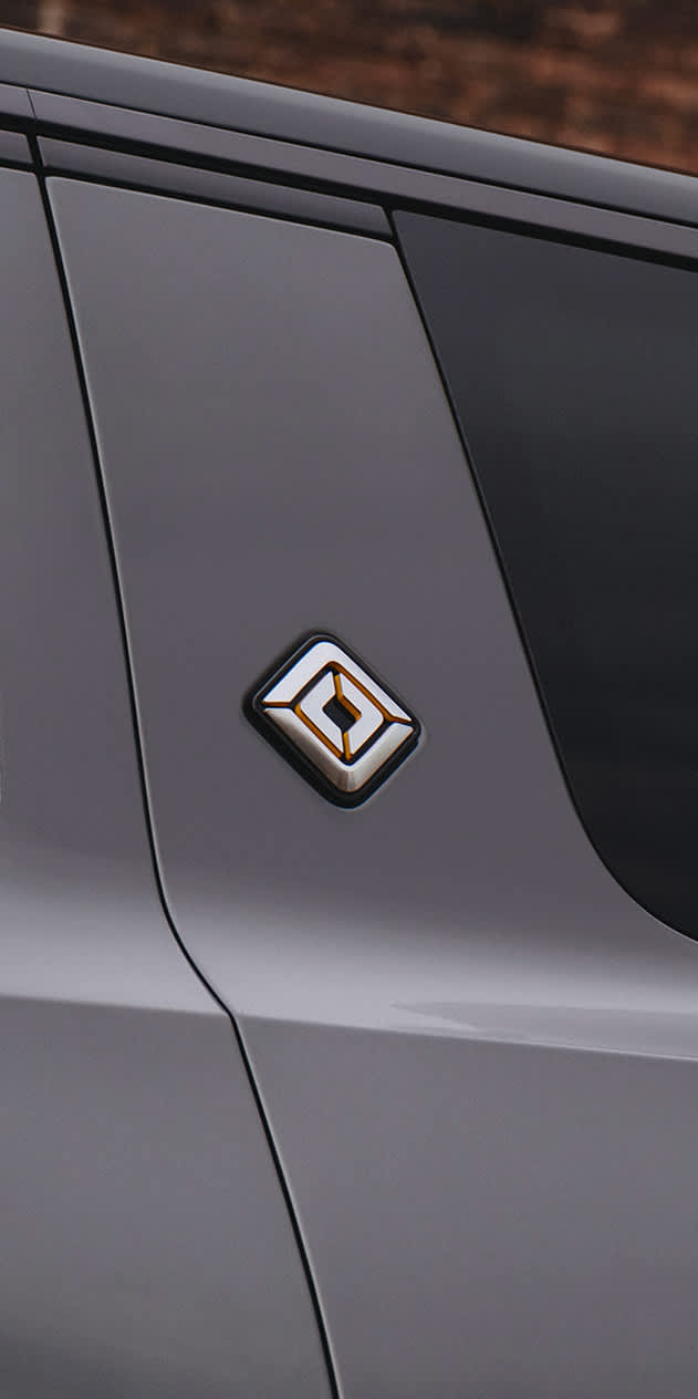 A side view of an electric Rivian R1S painted in Half Moon Grey is shown against a warm exposed brick wall.