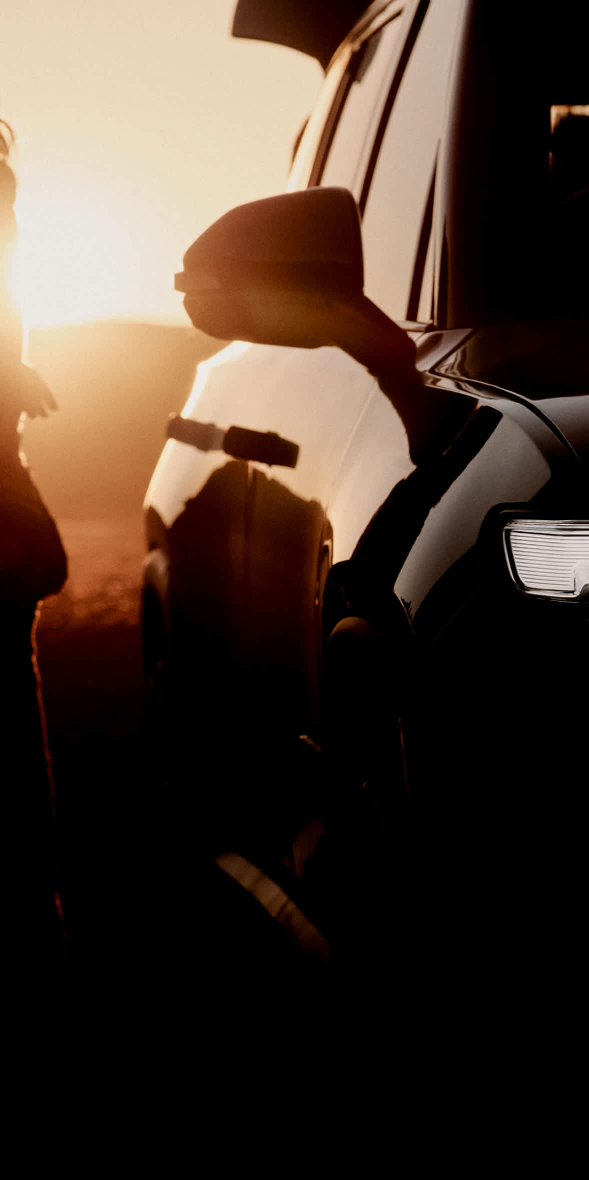 A person is seen next to a black Rivian R1S electric SUV with afternoon sun rays backlighting the scene