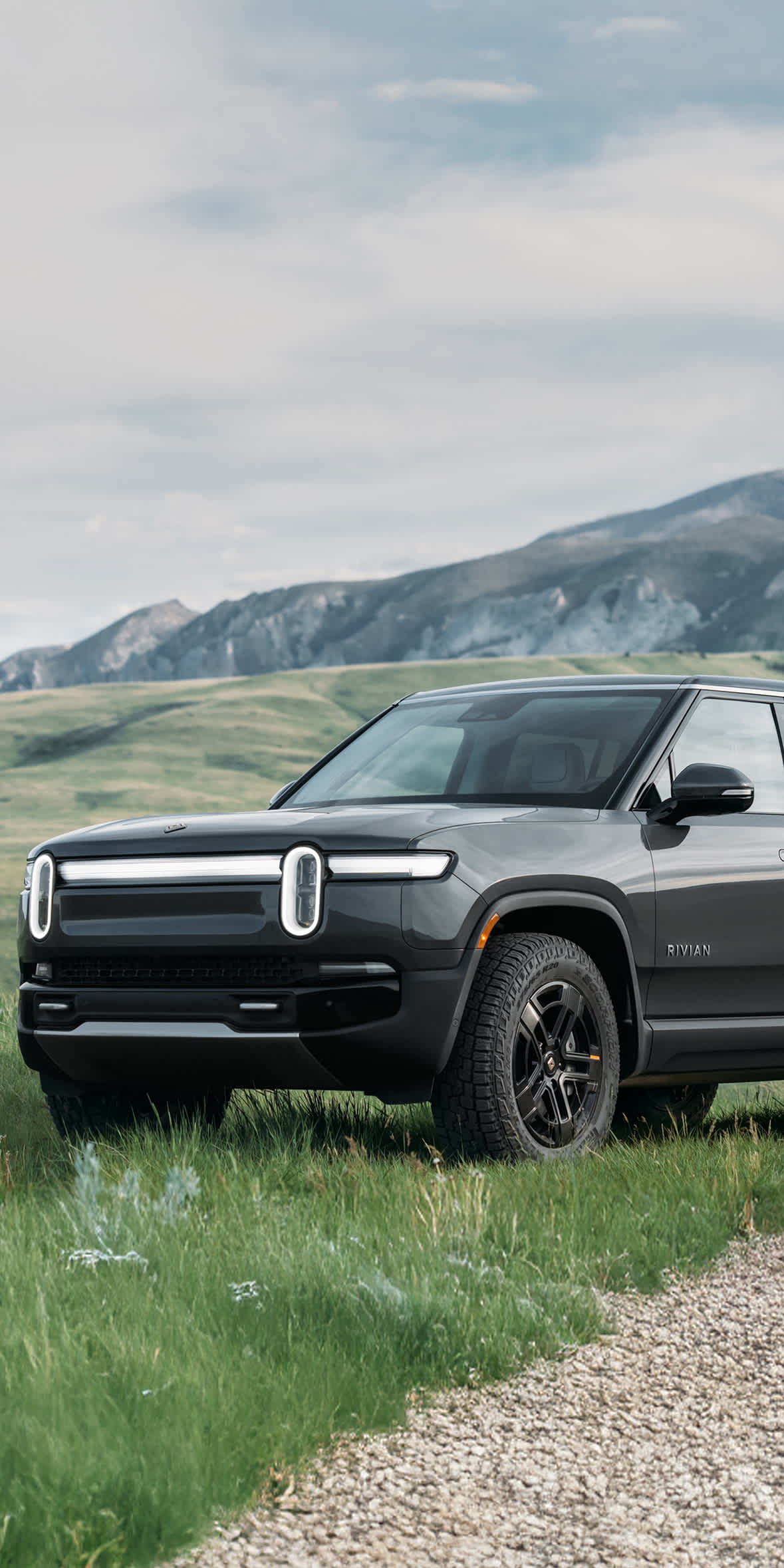 An image of a black Rivian R1S electric SUV parked in a grassy field with rolling hills and mountains in the background