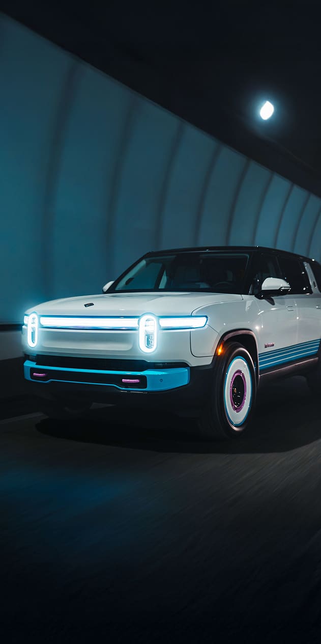 A side-front view of a Miami edition Rivian R1S electric SUV, white with bright blue and magenta accents, driving quickly through a dark tunnel. The signature light bar and headlights are brightly illuminated.