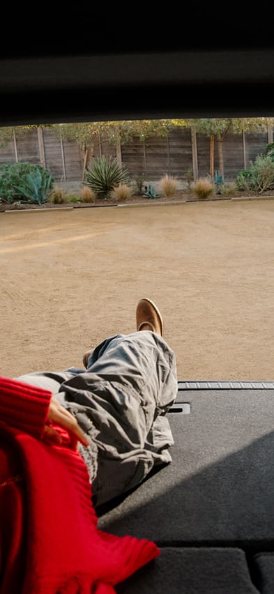 A person in a red sweater and grey trousers relaxes in the spacious back of R2 Rivian electric SUV with the rear hatch open