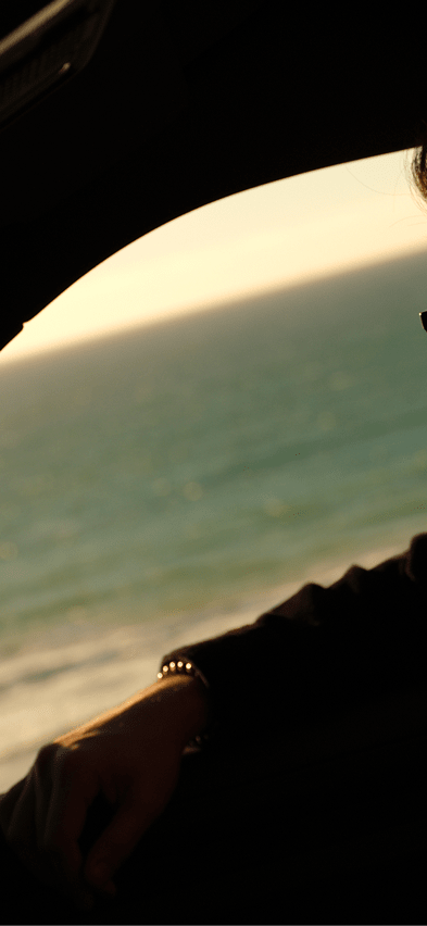 Silhouette of a man in sunglasses looking out the window of a Rivian R2 electric SUV at a golden ocean sunset along the coast.