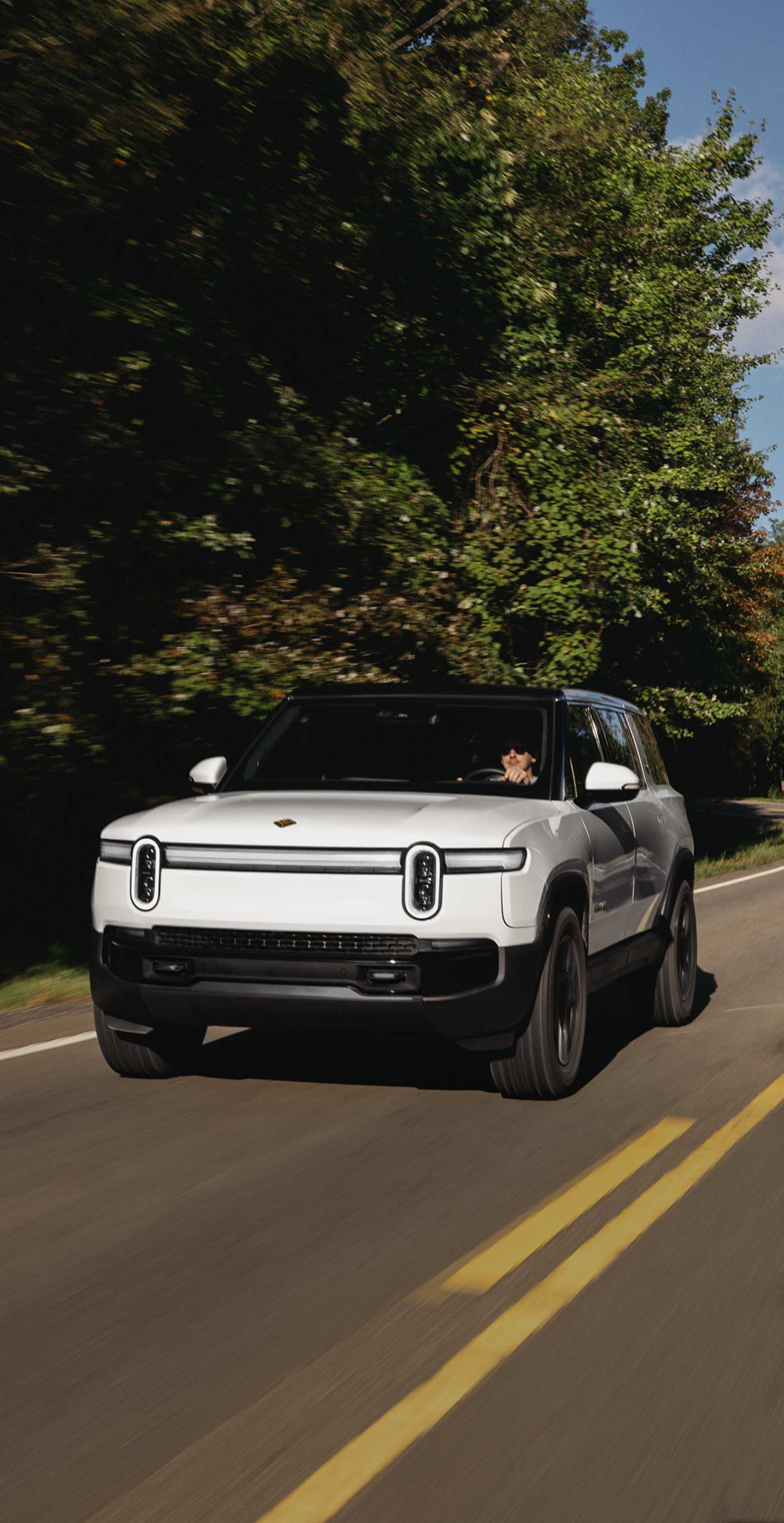A white Rivian R1S electric SUV driving swiftly down a two lane highway. Lush evergreen tress fill the background and line the sides of the road.