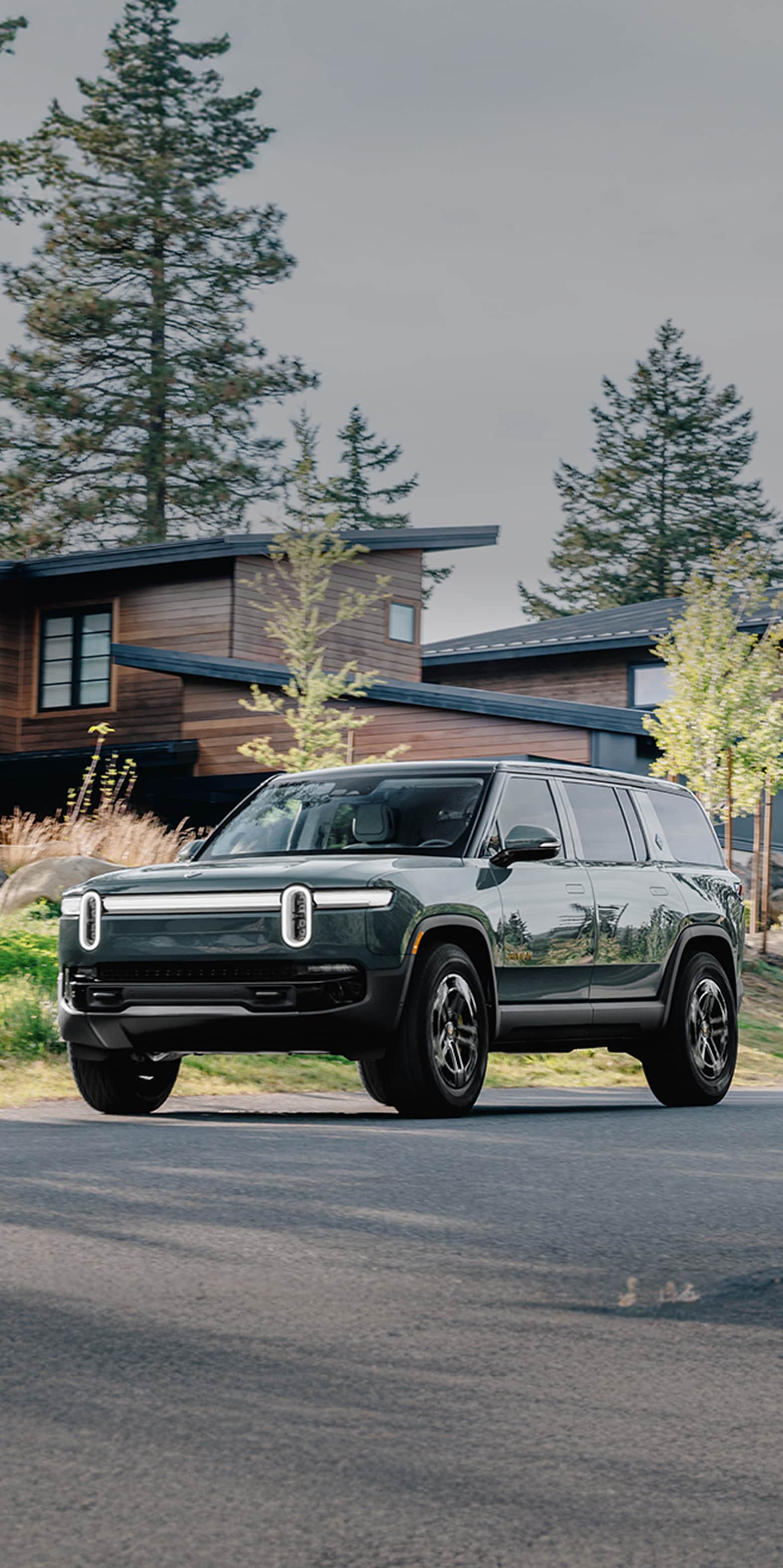 A side-front view of a Green Rivian R1S electric SUV traveling down a tree lined street in the fall. A modern house is in the background.
