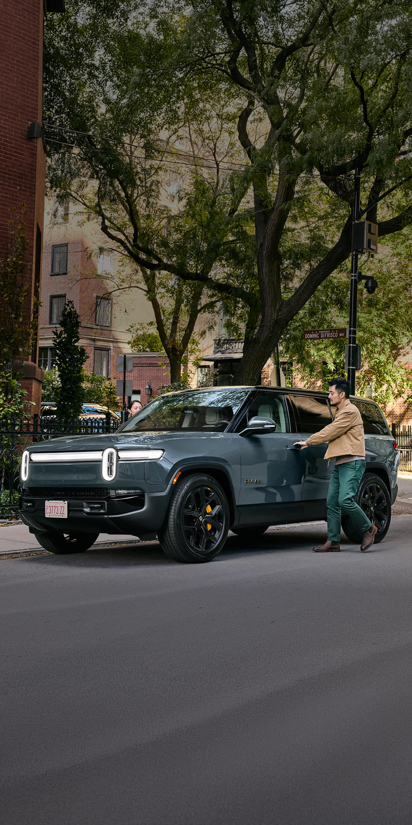 A man opening the driver door of a storm blue Rivian R1S electric SUV parked on a city street. A  green tree and picturesque brick apartments fill the background.