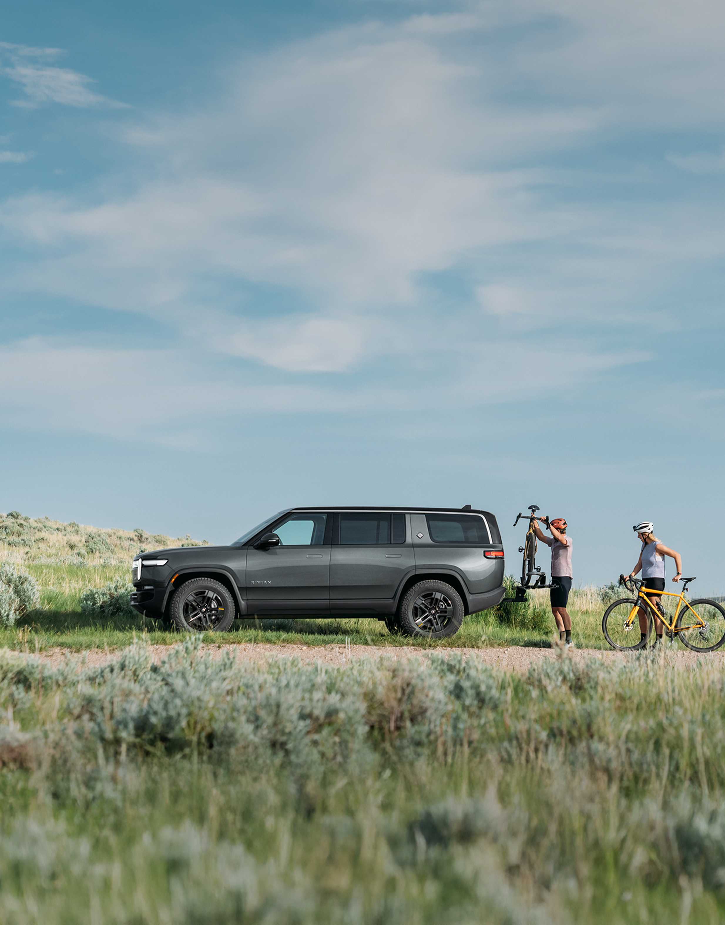 A white Rivian R1S electric SUV driving swiftly down a two lane highway. Lush evergreen tress fill the background and line the sides of the road.