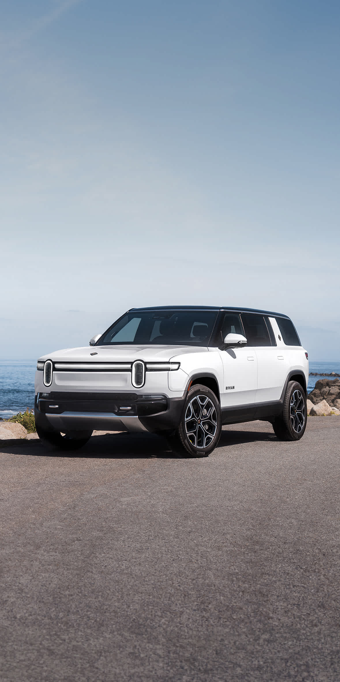 A white Rivian R1S electric SUV parked on an asphalt road next to a rocky ocean coastline under a clear sky.