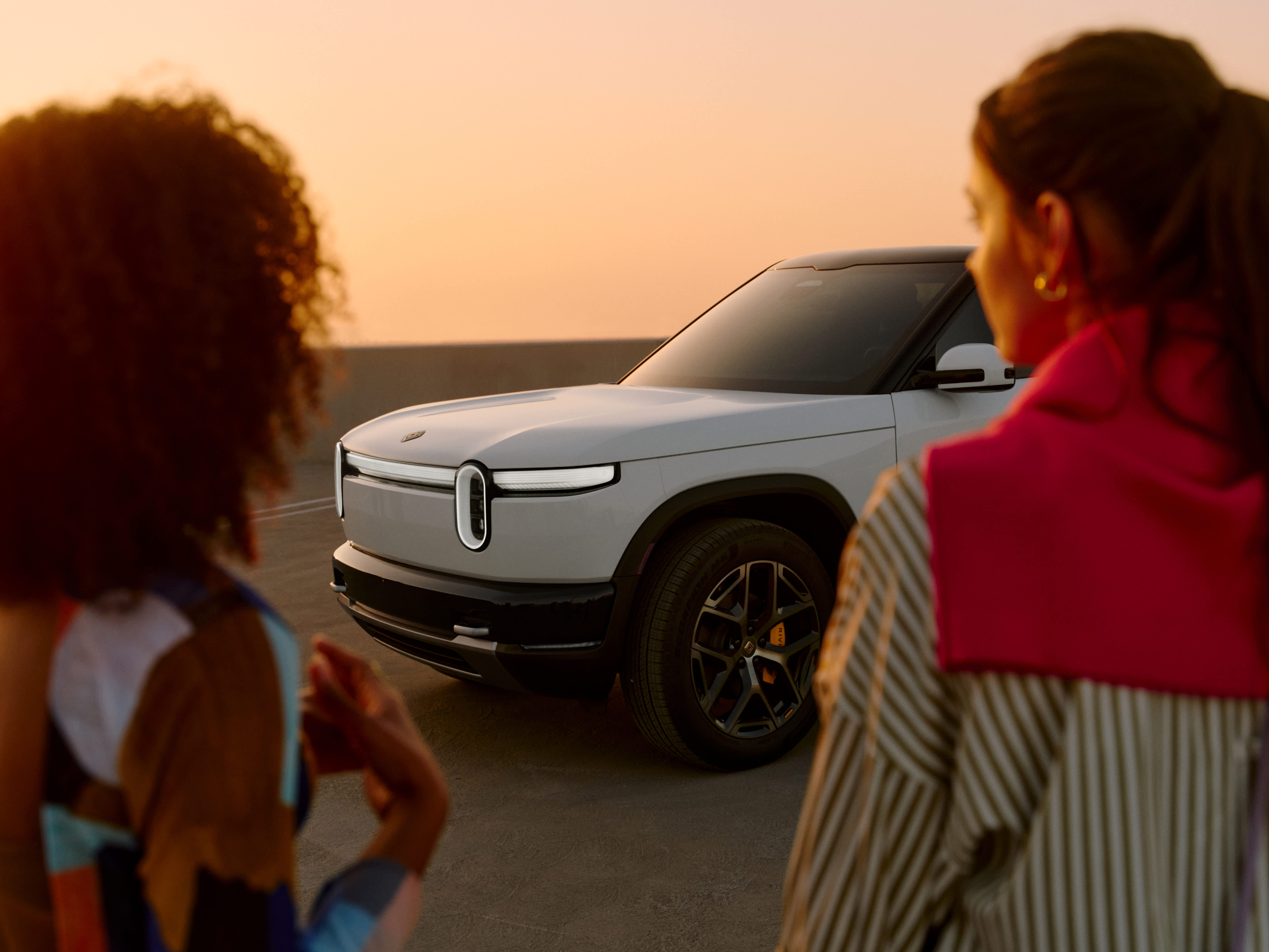 Two people seen from behind, looking at a white Rivian R2 SUV parked during a golden hour sunset.