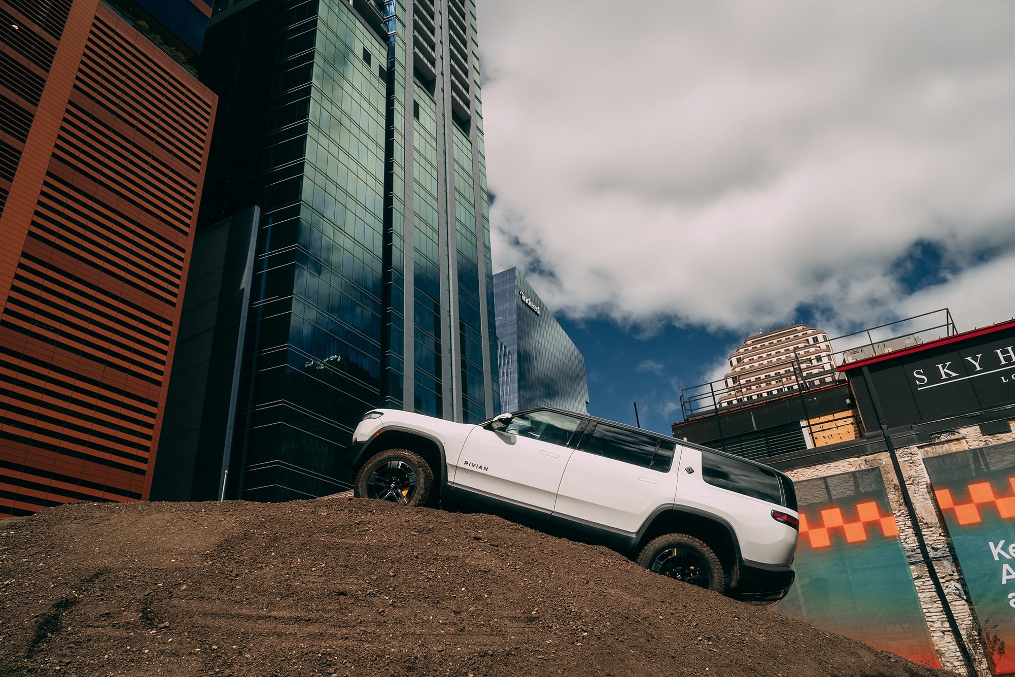 A white Rivian R1S SUV is parked at an angle on a large mound of dirt during the 2025 SXSW Electric Roadhouse. Behind the vehicle, towering modern glass skyscrapers rise into a blue sky filled with white clouds.