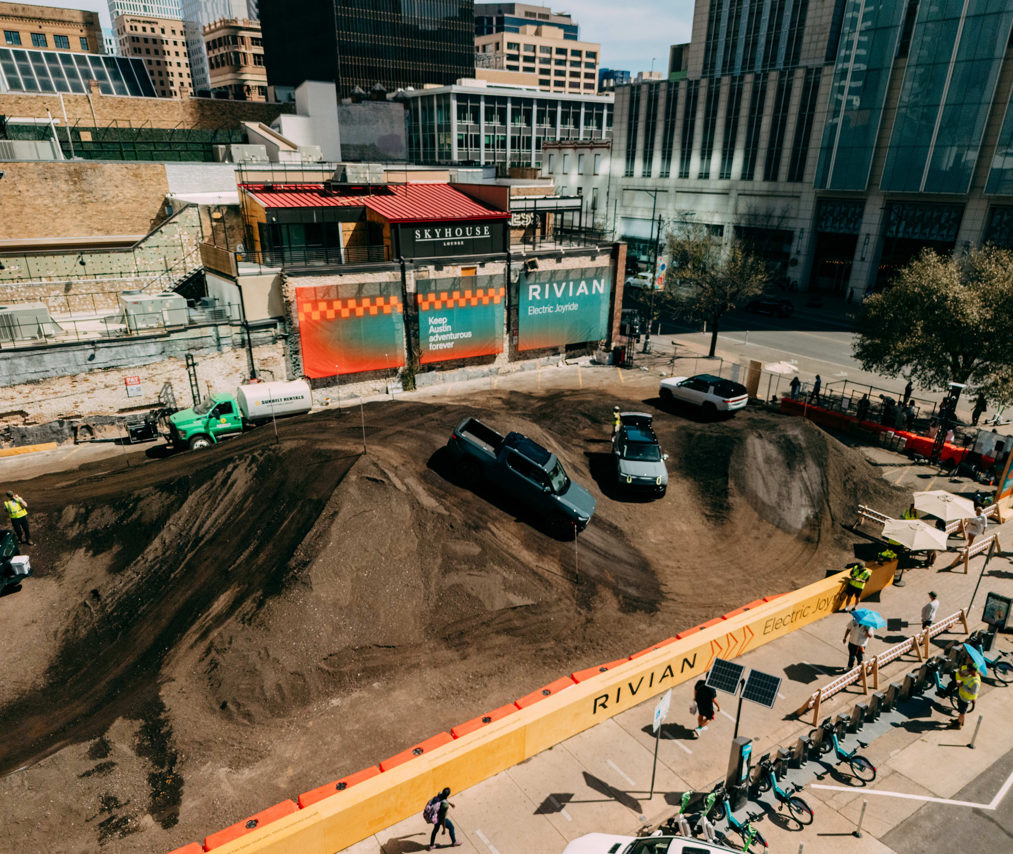 An aerial view of an urban dirt track course at the 2025 SXSW Electric Roadhouse, where multiple Rivian electric vehicles—including the R1T pickup and R1S SUV—are demonstrating their off-road capabilities