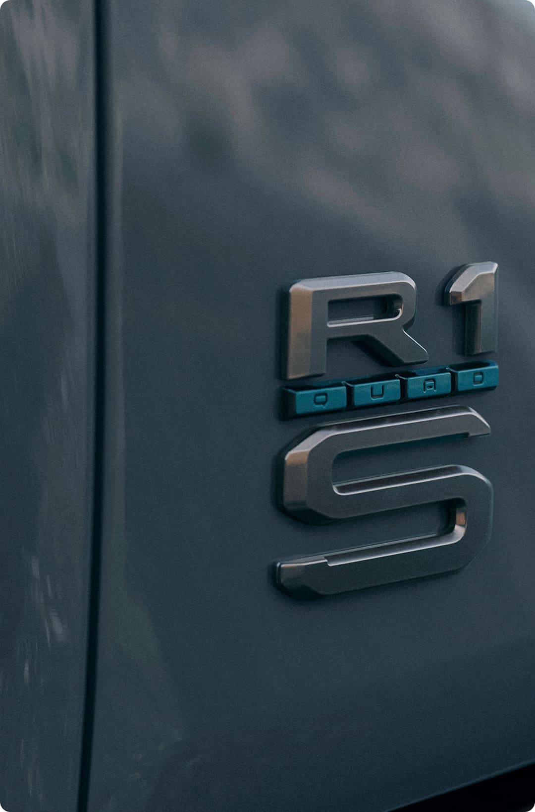Les badges arrière d'un véhicule électrique à quatre moteurs Rivian. Le badge montre le logo R1, mais comprend aussi un badge « QUAD » garni d'accents Bleu Laguna exclusifs au Quad.