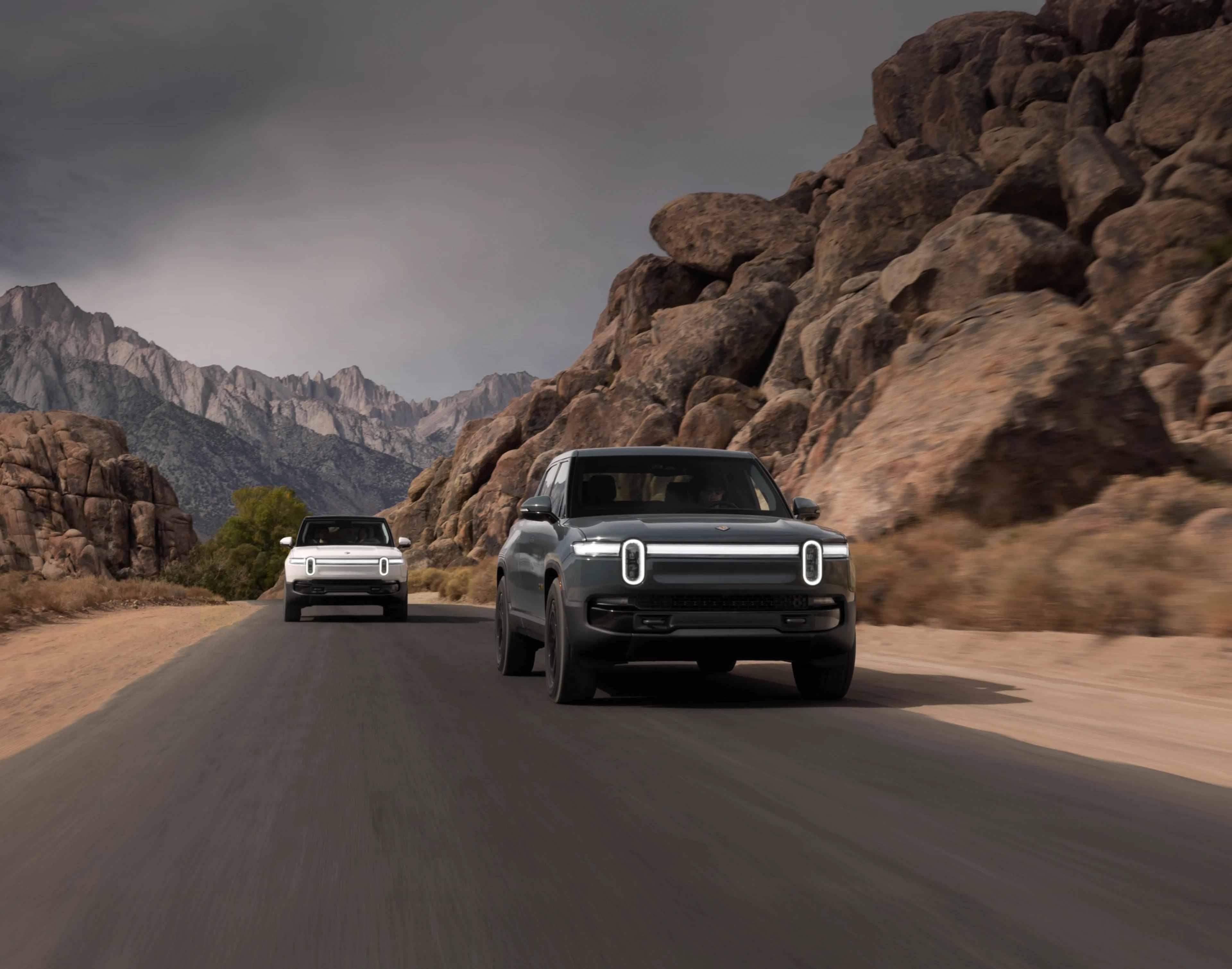 Deux VUS électriques Rivian R1S roulant sur une route de montagne pittoresque entourée de falaises rocheuses et d’un ciel nuageux.