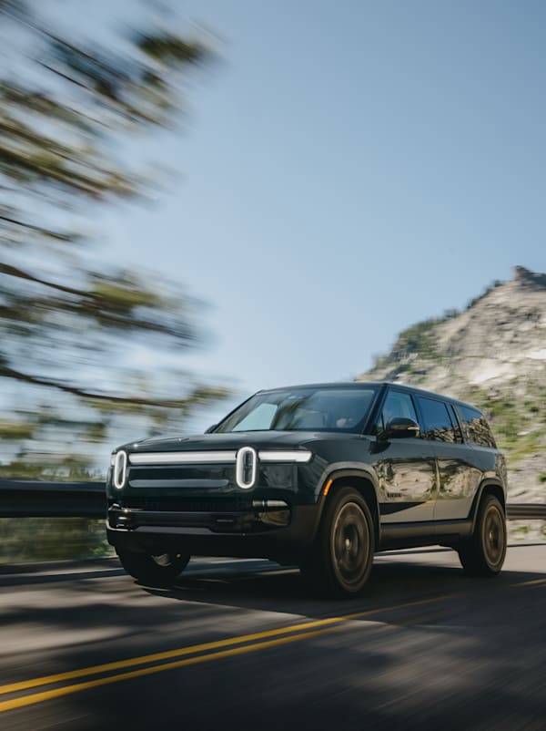 Un véhicule électrique à quatre moteurs Rivian Quad de couleur foncée. Ce VUS électrique est en mouvement sur une route bordée d'arbres avec une montagne en arrière-plan.
