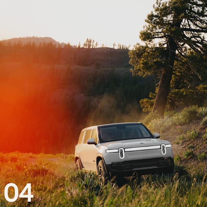 L'avant d'un VUS électrique à quatre moteurs Rivian R1S blanc roulant sur une route de terre avec de l'herbe, avec des collines arborées en arrière-plan.
