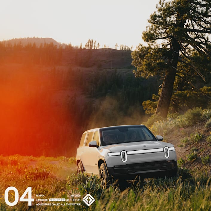 The front of a white Rivian R1S Quad motor electric SUV driving through grassy dirt road with tree-lined hills in the background.