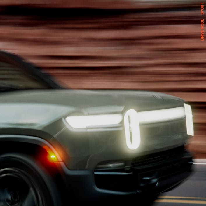 The driver side of a dark colored forest green Rivian R1S electric SUV. This quad motor vehicle shows motion blur as it moves down a small highway with redwood trees tree-filled hills in the background.