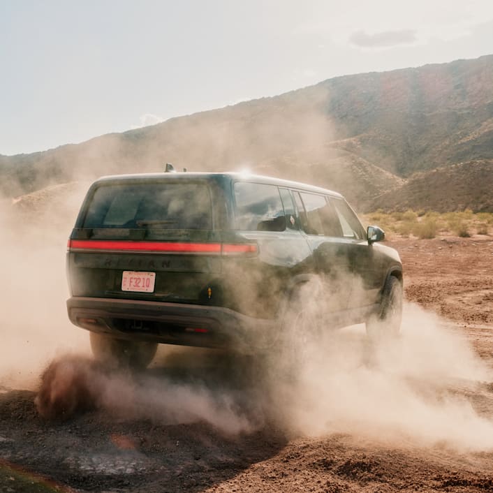 L'arrière d'un VUS électrique à quatre moteurs Rivian roulant dans une magnifique vallée en terre battue. La poussière se soulève et le véhicule fait un virage.