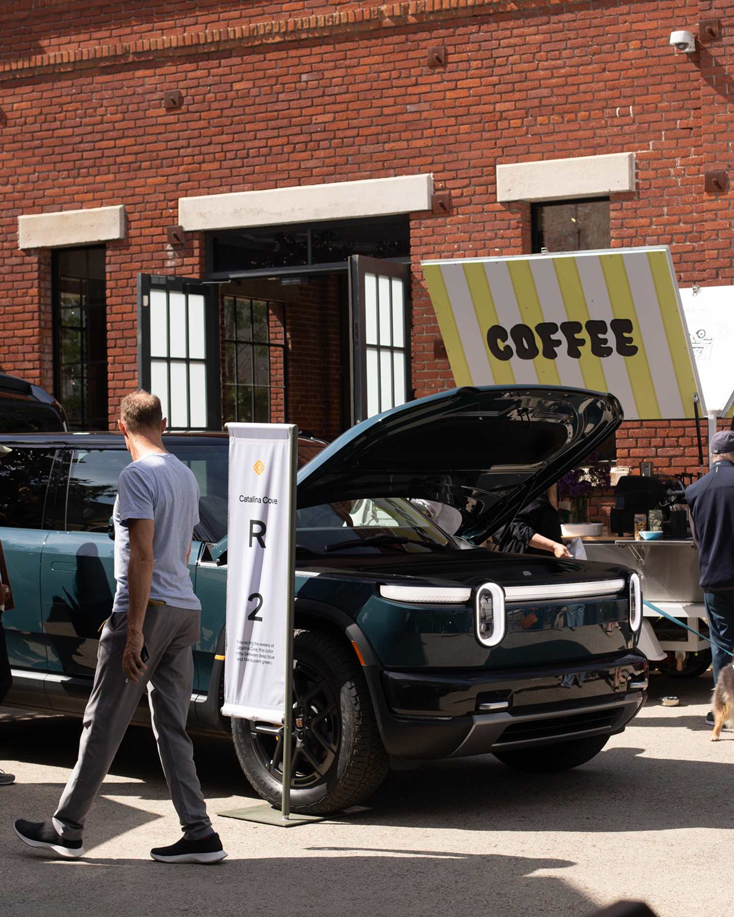 A Catalina Cove Rivian R2 with its hood open on display next to a coffee cart at a Rivian R2 Block Party.