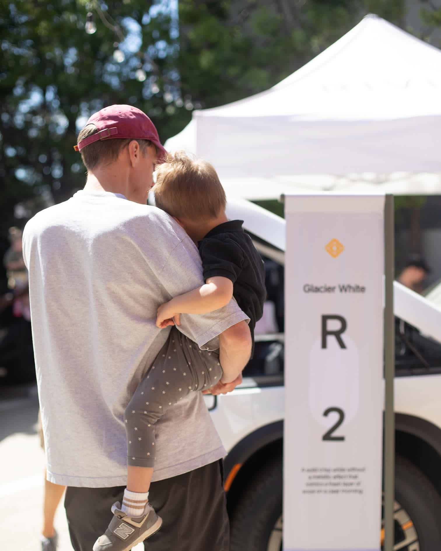 A father holding a toddler stands next to a "Glacier White R2" display banner at a Rivian R2 Block Party.